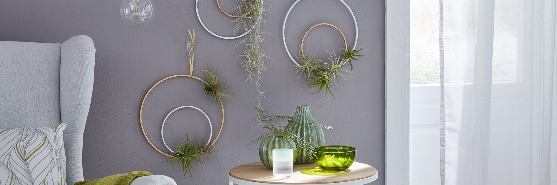 A modern living room corner features two round tables, one adorned with a green vase, glass bowl, and candle, while the other holds books. Air plants hang on circular wall art secured with tesa tape against a gray wall. An armchair with a green throw is positioned on the left. (This text has been generated by AI)