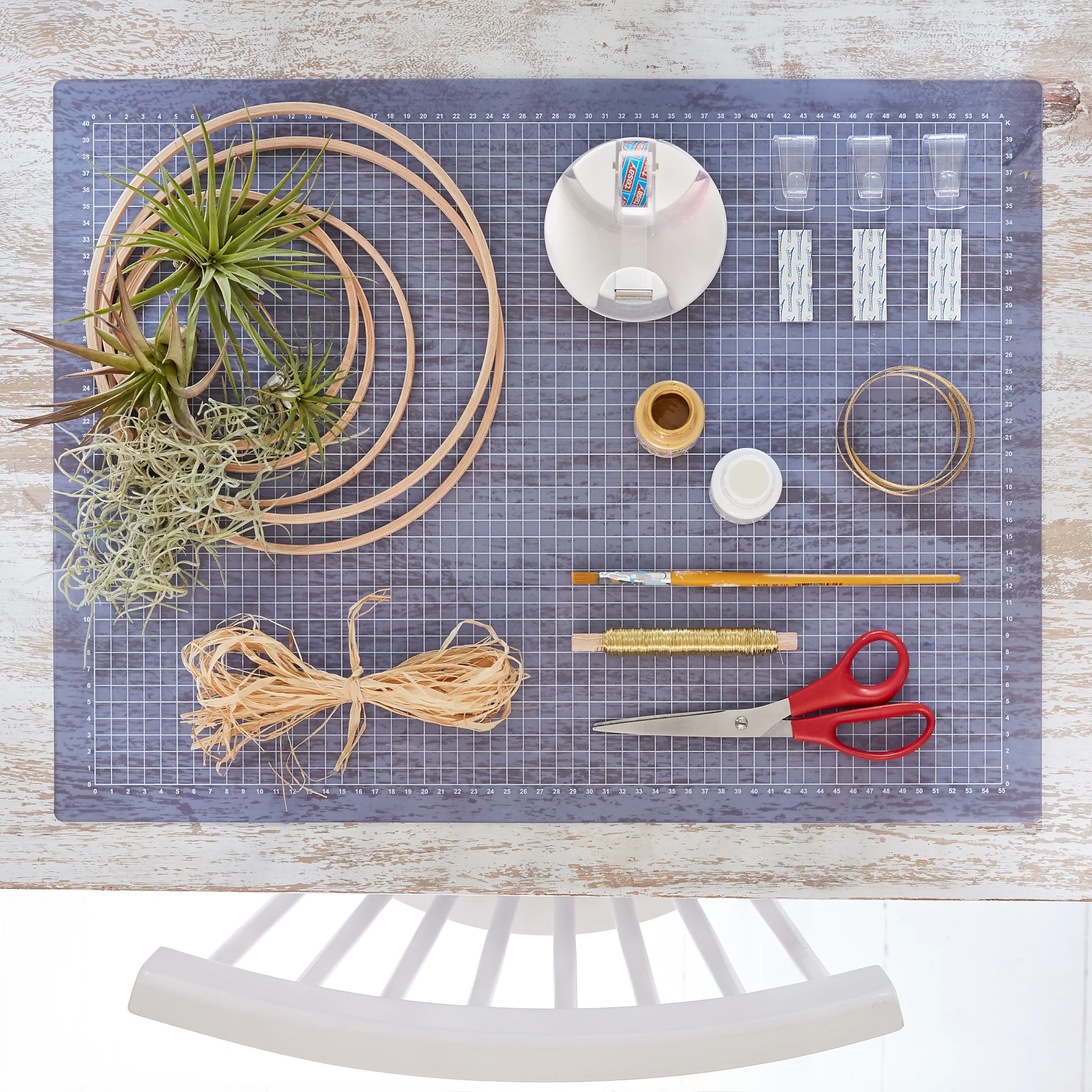 A wooden table holds crafting supplies: three air plants, embroidery hoops, tesa tape, thumbtacks, twine, string, a brush, scissors, and a cutting mat. A white chair is partially visible at the bottom. (This text has been generated by AI)