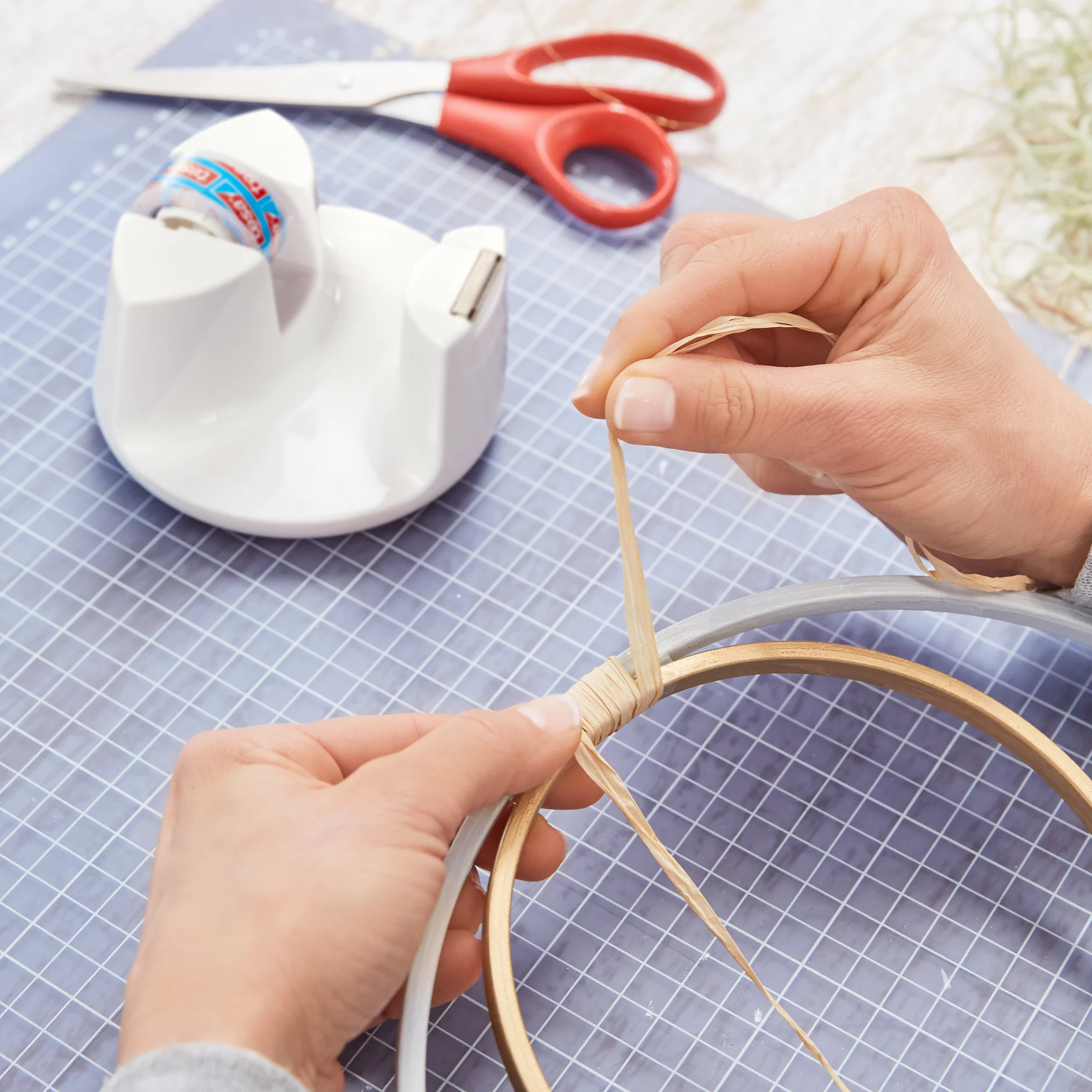 Two hands are wrapping a thin strip of material around a circular wooden frame. A roll of tesa tape and a pair of red scissors lie on top of a cutting mat in the background. A few thin strands are visible on the upper right corner of the image. (This text has been generated by AI)
