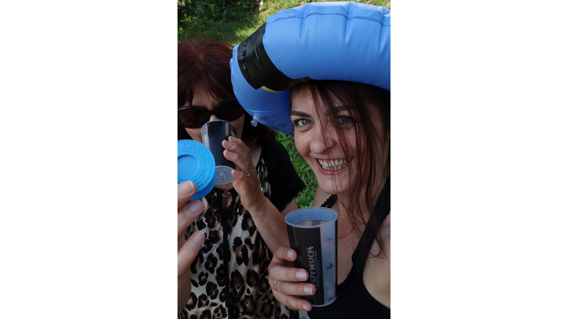 Zwei Frauen auf einer Wiese halten Plastikbecher. Eine Frau trägt eine Sonnenbrille und hält einen Deckel hoch, während die andere mit einem blauen aufblasbaren Ring auf dem Kopf in die Kamera lächelt. (This text has been generated by AI)