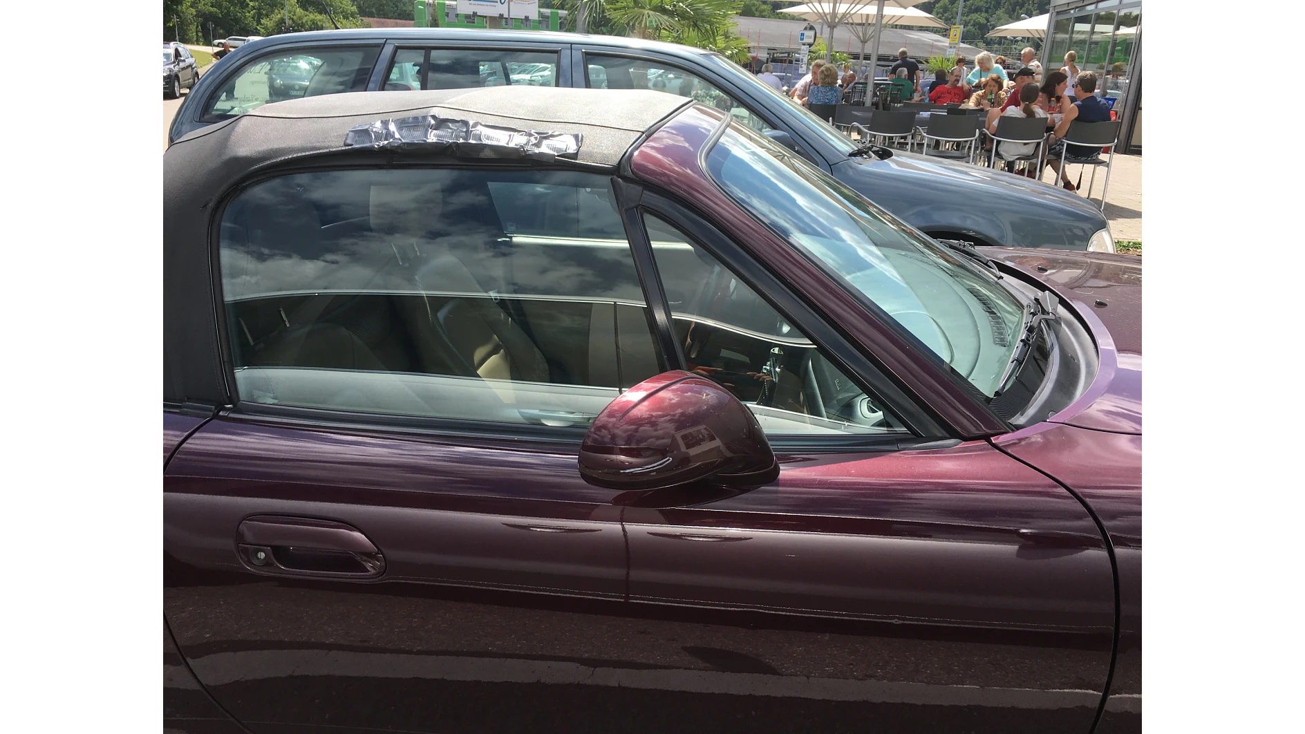 Ein kastanienbraunes Cabrio mit offenem Verdeck steht neben einem grauen Auto. Der Seitenspiegel reflektiert das Sonnenlicht. Im Hintergrund sitzen Menschen in ungezwungener Atmosphäre an Tischen im Freien unter Sonnenschirmen. (This text has been generated by AI)
