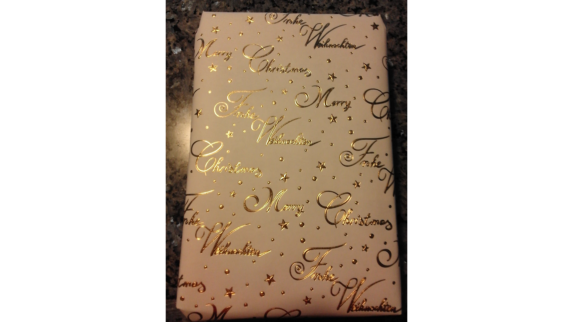 Ein verpacktes Geschenk in beigefarbenes Papier, verziert mit Goldfolienschriftzügen und kleinen Sternen, sicher gehalten durch Tesa-Klebeband. Die Worte „Merry Christmas“ und „Frohe Weihnachten“ sind auf der Verpackung wiederholt. Der Hintergrund ist eine dunkle, gesprenkelte Oberfläche. (This text has been generated by AI)