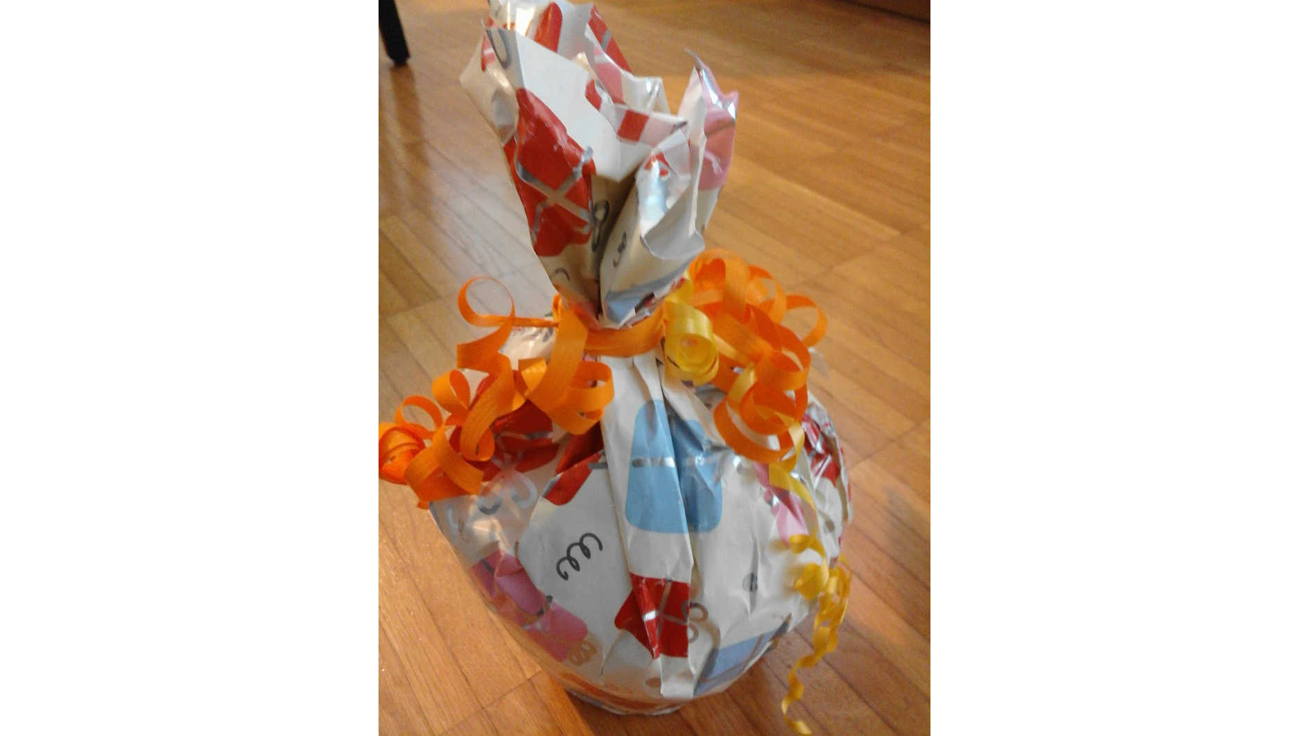 Ein Geschenk, eingewickelt in buntes Papier mit einem Muster aus Buchstaben und Formen. Es ist mit Tesa-Klebeband befestigt und mit gekräuselten orangefarbenen und gelben Bändern verschnürt. Das Geschenk liegt auf einem Holzboden. (This text has been generated by AI)