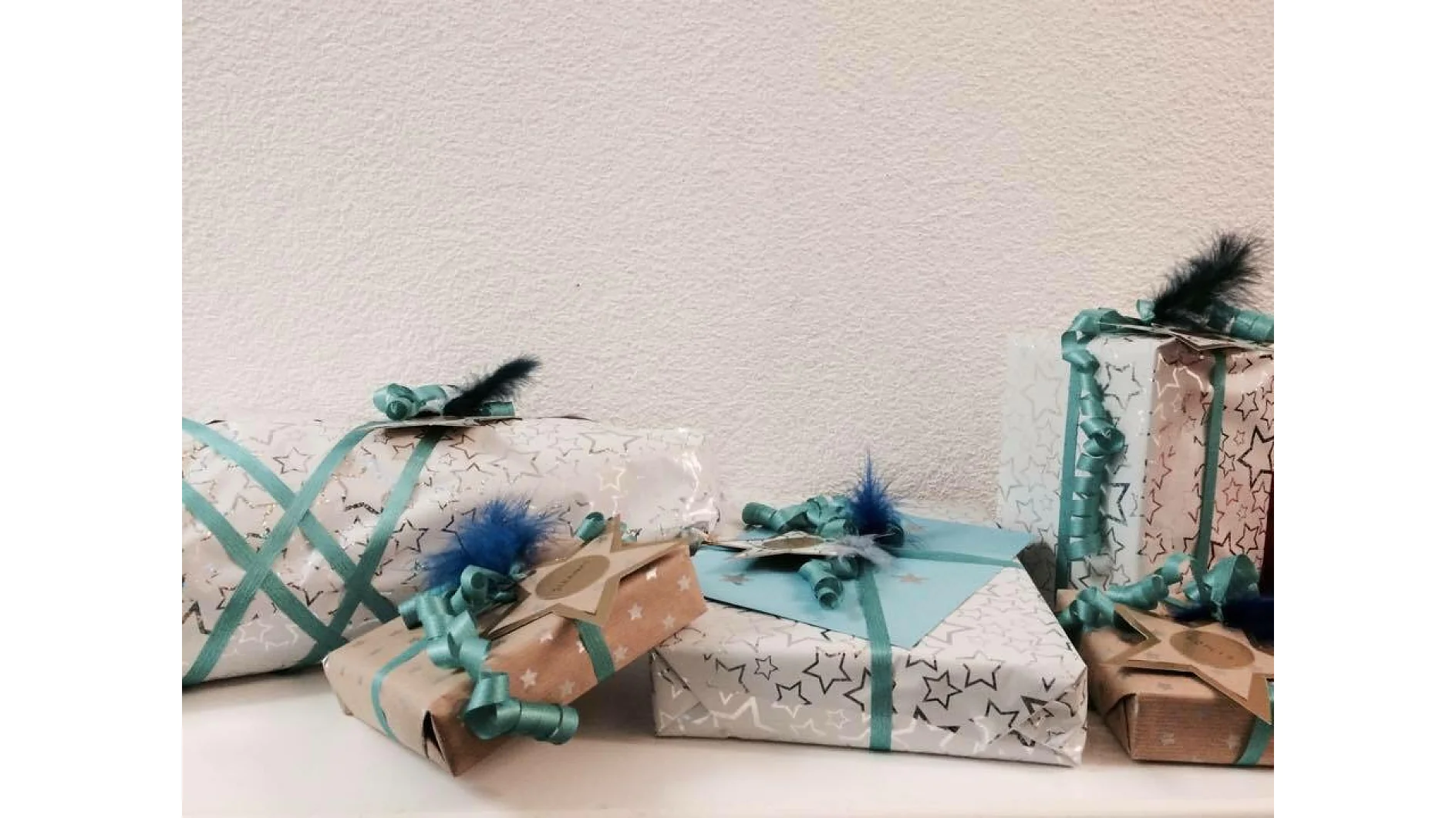 Fünf verpackte Geschenke sind auf einer Oberfläche vor einer strukturierten weißen Wand angeordnet. Die Geschenke sind mit sternenförmigem Papier mit blauen Bändern und Schleifen überzogen und alle mit Tesa-Klebeband befestigt. An einigen sind runde Etiketten mit Tesa-Klebeband befestigt und einige sind mit kleinen blauen Federn geschmückt, die mit Tesa-Klebeband an ihrem Platz gehalten werden. (This text has been generated by AI)