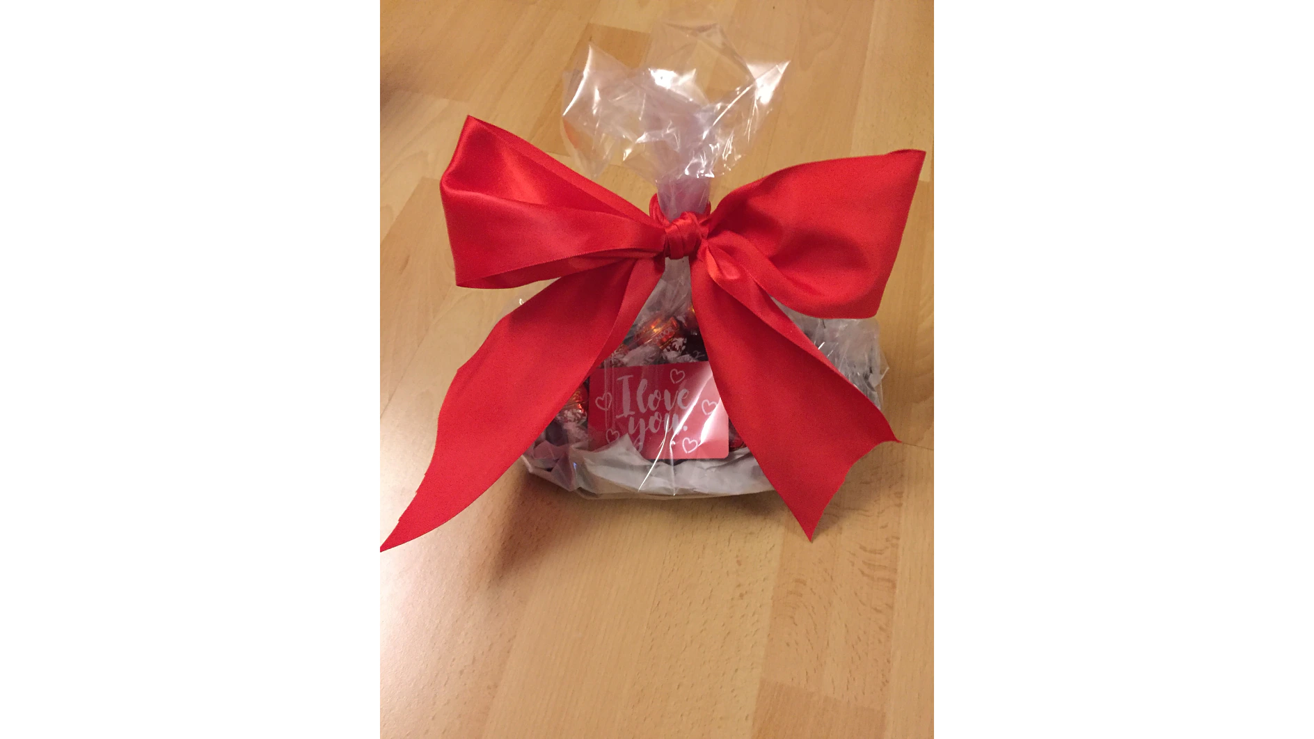 Ein Geschenkkorb ist in durchsichtige Folie eingewickelt und mit einer großen roten Schleife zusammengebunden. Darin befinden sich verschiedene kleine Geschenke, darunter Pralinen und eine Karte mit der Aufschrift „Ich liebe dich“ auf rosa Hintergrund mit Herzmotiven. Die Folie ist mit Tesa-Klebeband sicher befestigt, damit alles schön verpackt bleibt. Der Korb steht auf einem Holzboden. (This text has been generated by AI)