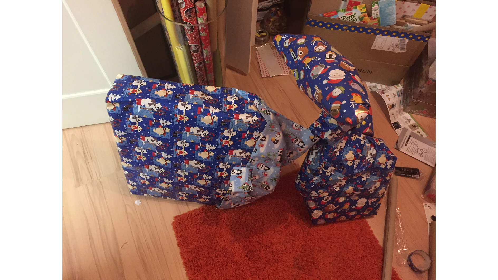 Drei seltsam geformte Geschenke, die in festliches Papier mit Cartoon-Motiven eingewickelt sind, liegen auf einem Holzboden. Im Hintergrund sind einige Geschenkverpackungen zu sehen. Die Geschenke liegen auf einem orangefarbenen Teppich neben Geschenkpapierrollen und einem Karton. Das Tesa-Klebeband, mit dem die Geschenke verschlossen sind, verleiht ihnen ein hübsches Finish und trägt zu ihrem verspielten Aussehen bei. (This text has been generated by AI)