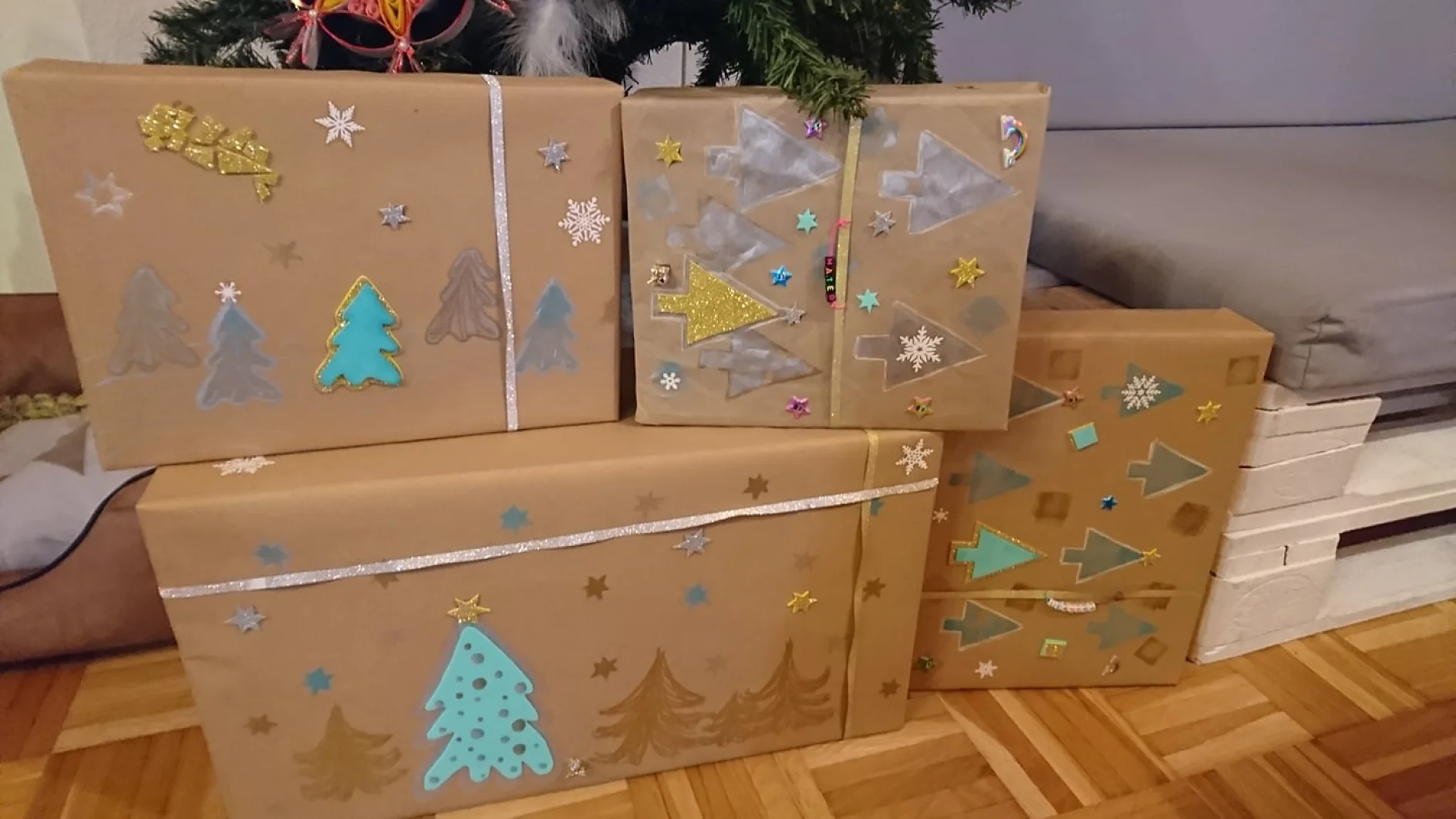 Mehrere verpackte Geschenke liegen auf einem Holzboden neben einem Kissen und einem Baum. Die Pakete sind mit braunem Papier überzogen und mit bunten Papierausschnitten von Bäumen, Sternen und Schneeflocken verziert. An einer der Schachteln befindet sich eine silberne Schleife, die mit Tesa-Klebeband sicher befestigt ist. (This text has been generated by AI)