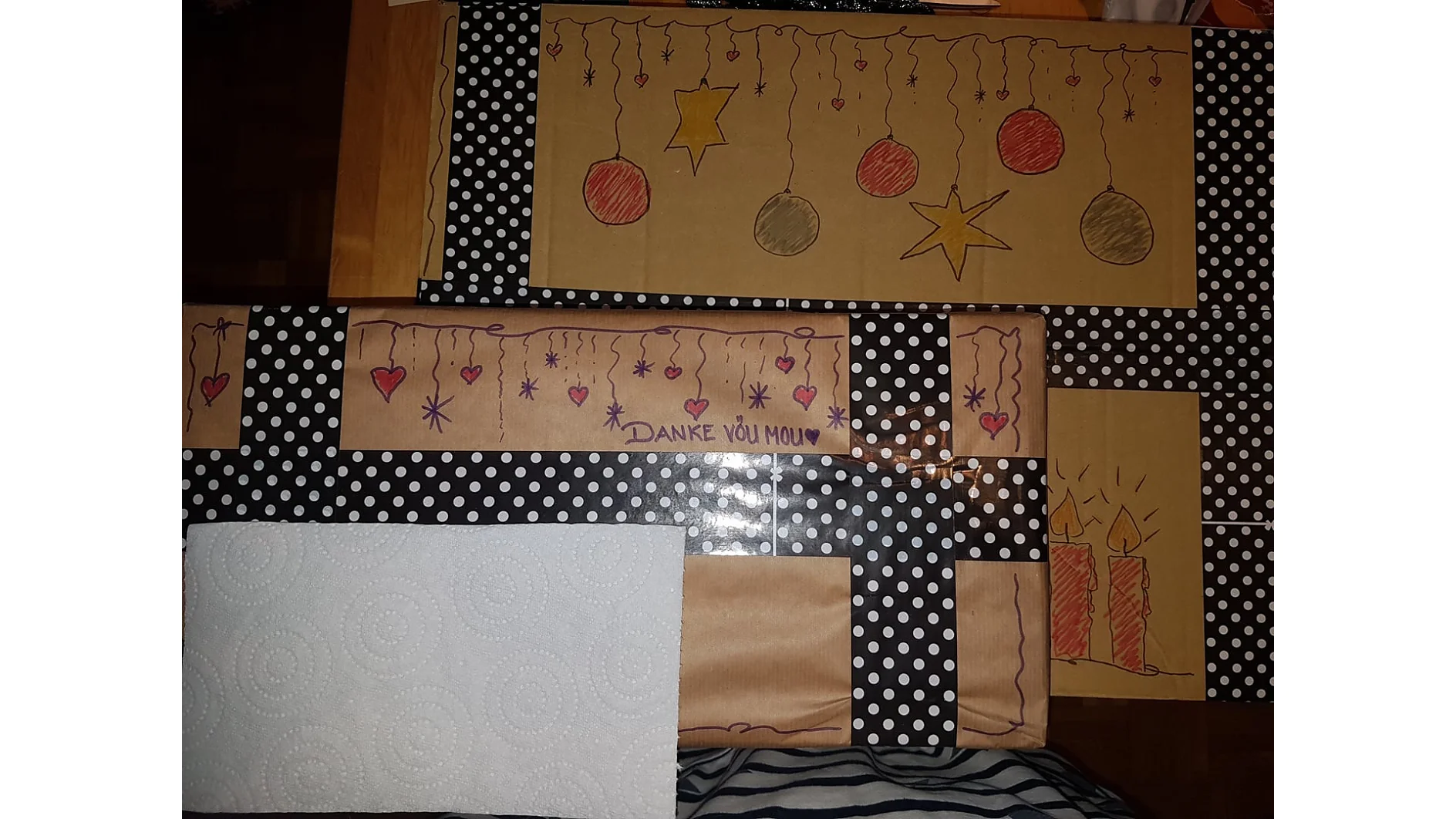Geschenke, die in braunes Papier mit handgemalten Weihnachtsdekorationen wie Sternen und Kugeln eingewickelt sind, werden mit Tesa-Klebeband zusammengehalten. Zusätzlich gibt es schwarz-weißes Geschenkpapier mit Punkten, das mit Tesa-Klebeband zusammengehalten wird. Ein Papiertuch bedeckt die Geschenke teilweise. Die Worte „Danke You Mou“ sind sichtbar. (This text has been generated by AI)