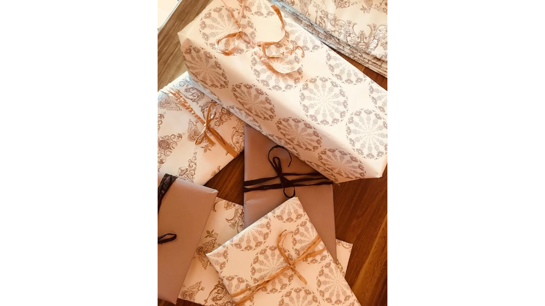 Mehrere verpackte Geschenke sind auf einer Holzoberfläche gestapelt. Sie sind mit gemustertem Papier mit kreisförmigen Motiven überzogen und mit Tesa-Klebeband befestigt, während sie mit Schnüren in Braun und Schwarz zusammengebunden sind. Die Geschenke variieren in Größe und Form. (This text has been generated by AI)
