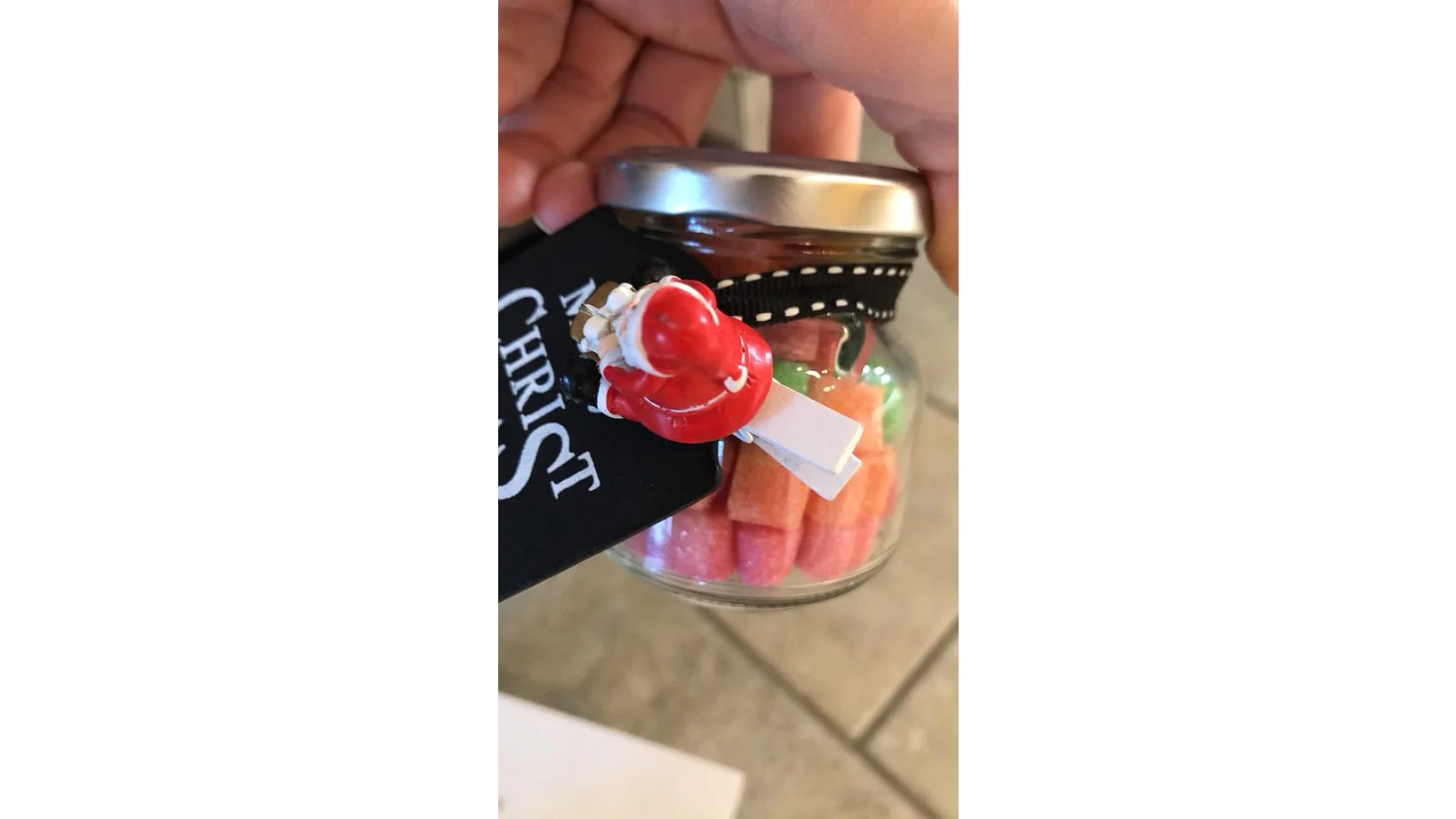 Ein kleines Glasgefäß mit silbernem Deckel ist mit rosa, orangefarbenen und grünen Süßigkeiten gefüllt. An der Seite ist mit Tesa-Klebeband und schwarzem Band eine rote Figur befestigt, die an den Weihnachtsmann erinnert. An dem Glas ist ein schwarzes Etikett mit teilweise sichtbarem weißem Text befestigt. (This text has been generated by AI)