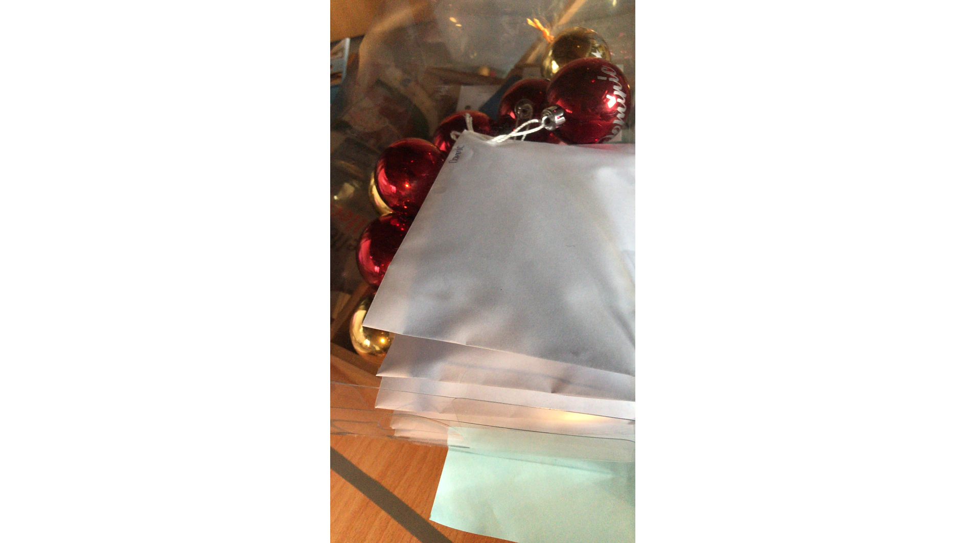 Ein Stapel Papier bedeckt teilweise eine Ansammlung roter und goldener Weihnachtsdekorationen. Die Objekte sind auf einer Holzoberfläche platziert und die Szene scheint sich in einem Innenbereich abzuspielen. (This text has been generated by AI)