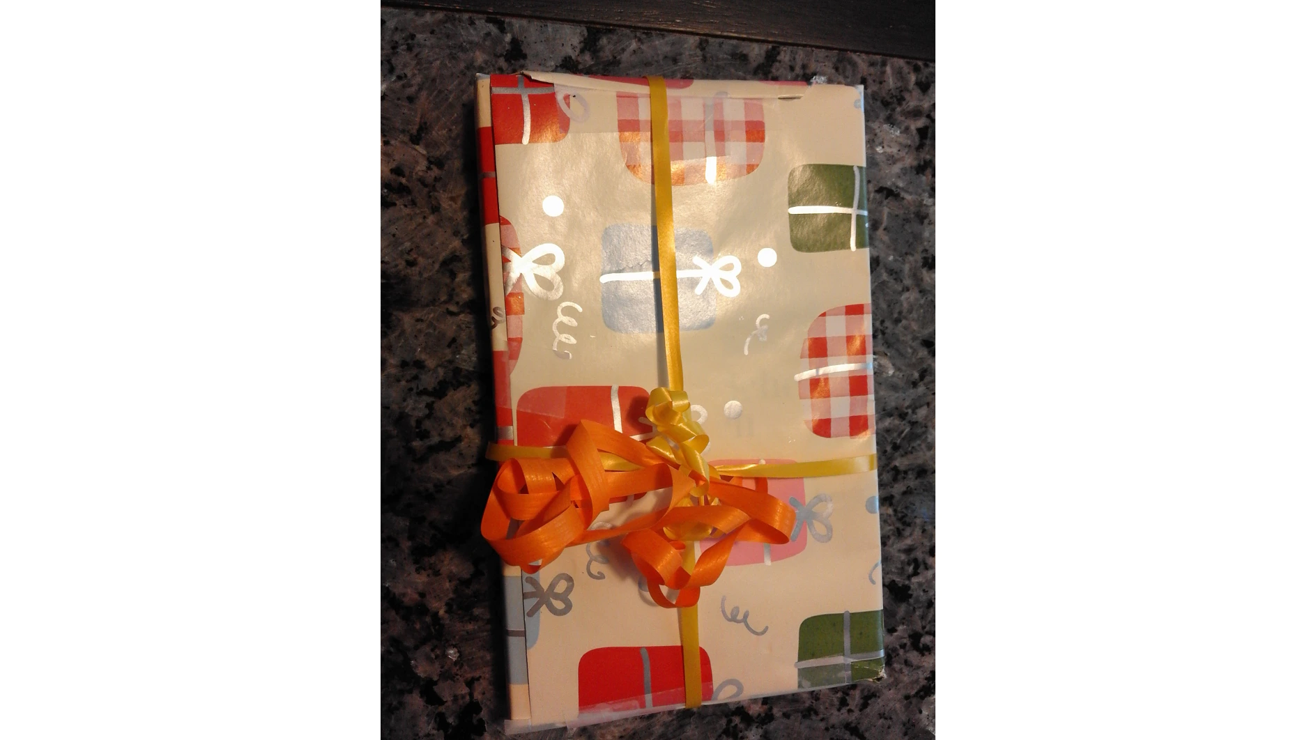 Ein Geschenk, eingewickelt in buntes Papier mit einem Muster aus roten, grünen und orangefarbenen Geschenken. Es ist mit einem gelben Band und einer Schleife aus Tesa-Klebeband zusammengebunden. Das Geschenk wird auf einer Granitarbeitsplatte platziert. (This text has been generated by AI)