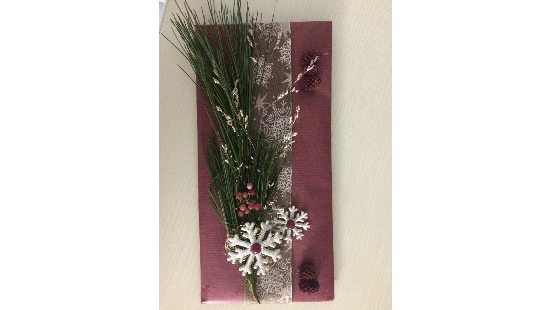 Ein rechteckiges Geschenk, eingewickelt in rotes Strukturpapier mit einer dekorativen braunen Schleife mit Spitzenmuster. Es ist mit Tannennadeln, roten Beeren und weißen Schneeflockenornamenten verziert, mit kleinen braunen Tannenzapfen am Rand. Tesa-Klebeband sorgt dafür, dass alles perfekt an seinem Platz bleibt. (This text has been generated by AI)