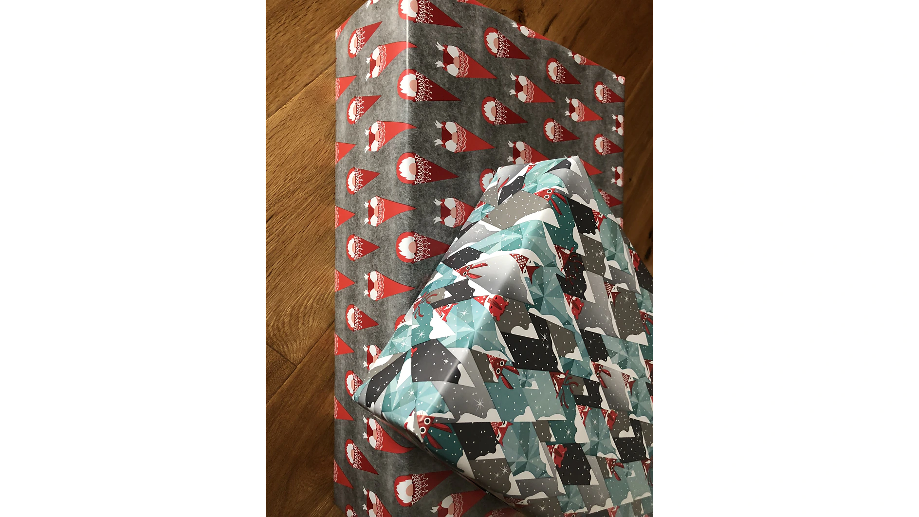 Auf einem Holzboden liegen zwei verpackte Geschenke. Ein Geschenk ist in rotes Papier mit Zwergenmotiven eingewickelt und sorgfältig mit Tesa-Klebeband befestigt. Das andere Geschenk ist mit Papier mit einem Muster aus abstrakten Formen in Blau, Schwarz und Weiß mit kleinen Fuchsillustrationen verziert und ebenfalls fachmännisch mit Tesa-Klebeband verschlossen. (This text has been generated by AI)