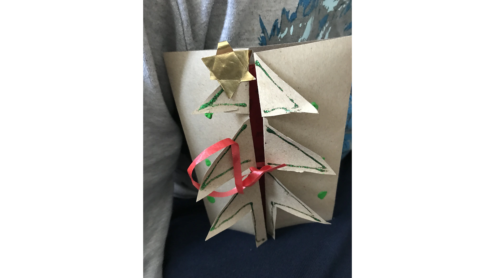 Eine handgefertigte Karte mit einem Pop-up-Weihnachtsbaum-Design. Der Baum ist mit grünem Glitzer verziert und mit einem goldenen Stern gekrönt. Ein rotes Band ist um die Karte gewickelt und mit Tesa-Klebeband befestigt. Die Karte besteht aus braunem Papier, sodass alles schön an seinem Platz bleibt. (This text has been generated by AI)