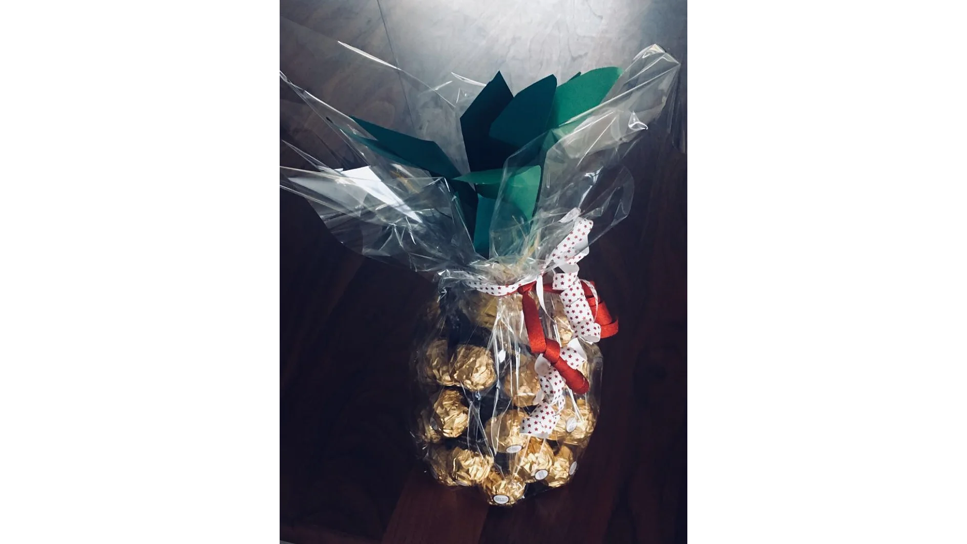 Ein Geschenkarrangement in Ananasform mit einzeln verpackten Pralinen aus Goldfolie und grünen Papierblättern. Es ist in durchsichtiges Zellophan eingewickelt, mit Tesa-Klebeband befestigt und mit einer weiß-roten Schleife zusammengebunden. (This text has been generated by AI)
