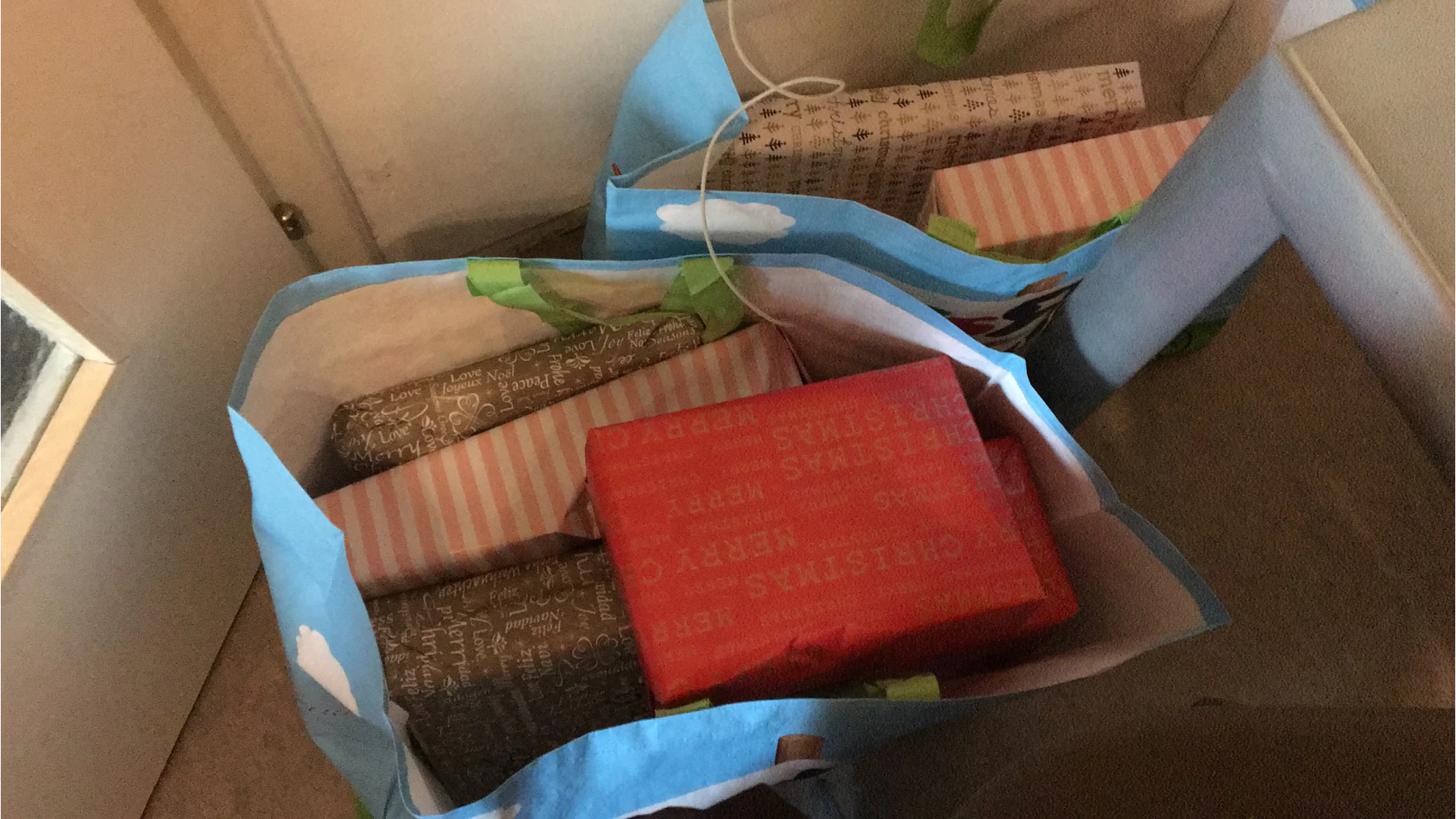 Drei blaue Geschenktüten sind mit verschiedenen Geschenken gefüllt, die mit Tesa-Klebeband verpackt sind. Das Geschenkpapier ist rot, grau mit weißem Text und rosa Streifen. Die Tüten liegen nebeneinander auf einem mit Teppich ausgelegten Boden. (This text has been generated by AI)