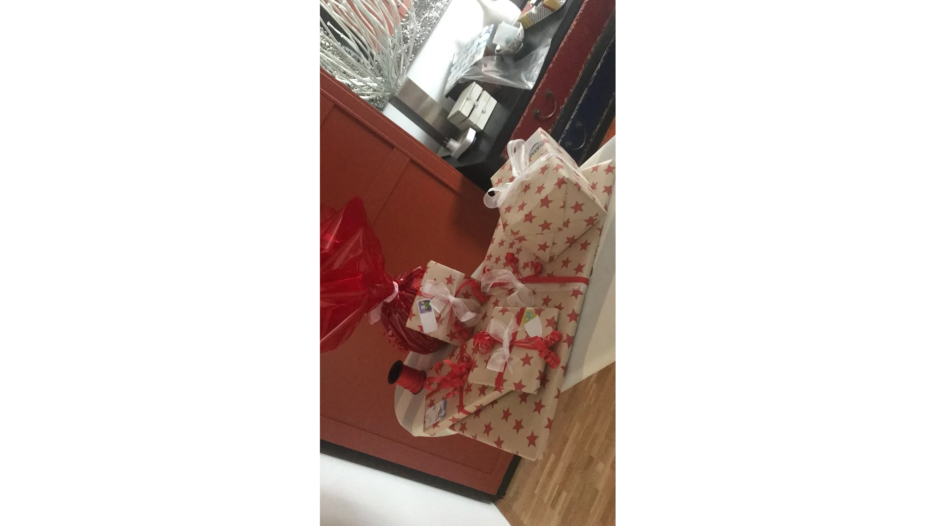 Auf einer weißen Fläche sind verschiedene verpackte Geschenke angeordnet. Das größte Paket ist in weißes Papier mit roten Sternen eingewickelt und mit Tesa-Klebeband befestigt. Andere Geschenke sind mit roten Bändern geschmückt und mit Tesa-Klebeband befestigt, und eines ist mit Tesa-Klebeband in rotes Zellophan eingewickelt. An jedem Geschenk sind Etiketten befestigt. (This text has been generated by AI)