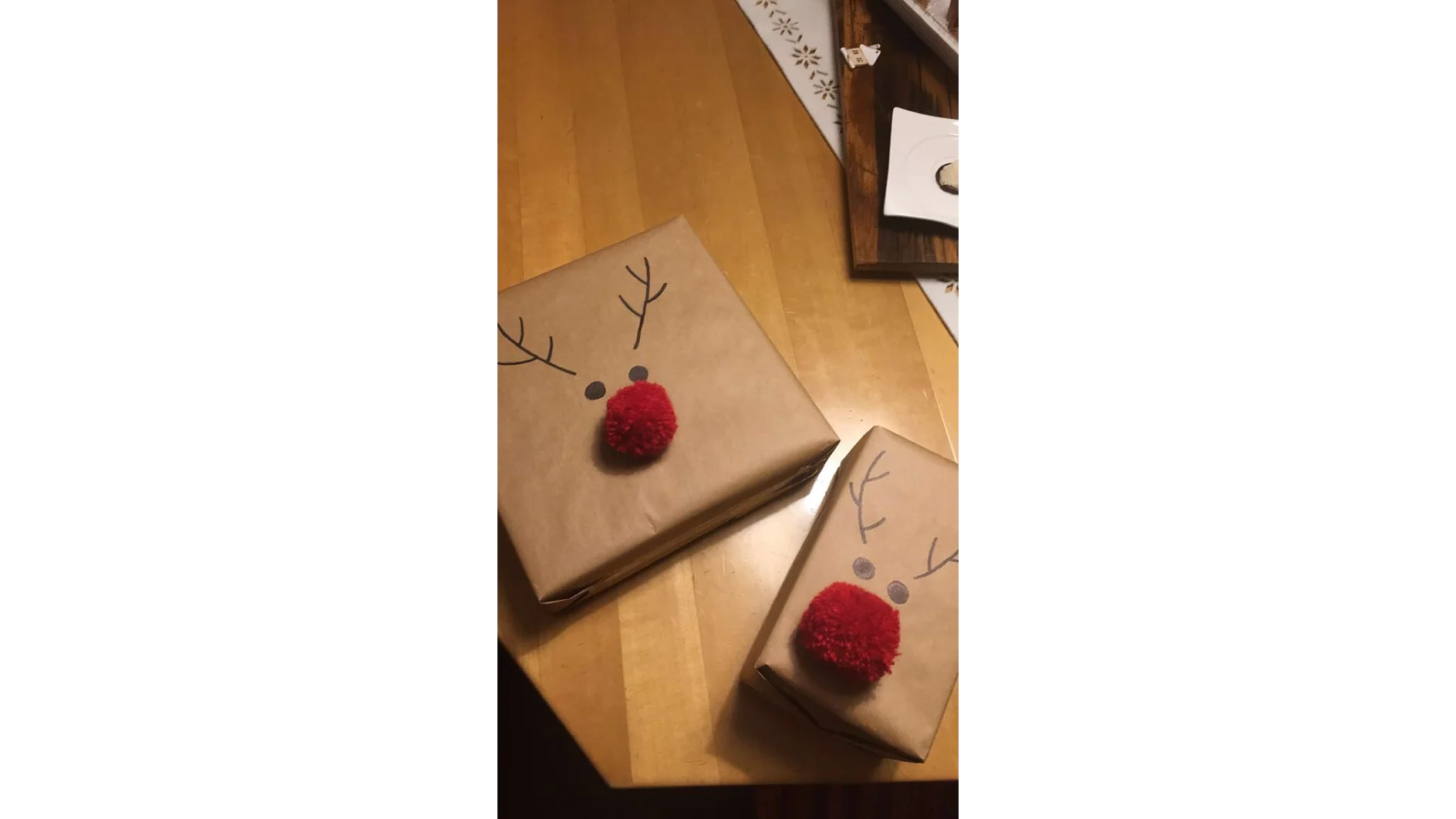 Auf einem Holztisch liegen zwei in braunes Papier eingewickelte Geschenke mit Rentiermotiven. Jedes hat einen roten Pompon als Nase und schwarze Linien als Geweih, alles mit Tesaband befestigt. Ein Geschenk ist größer als das andere. Im Hintergrund sind eine teilweise sichtbare Deckchenspitze und ein Holzbrett zu sehen. (This text has been generated by AI)