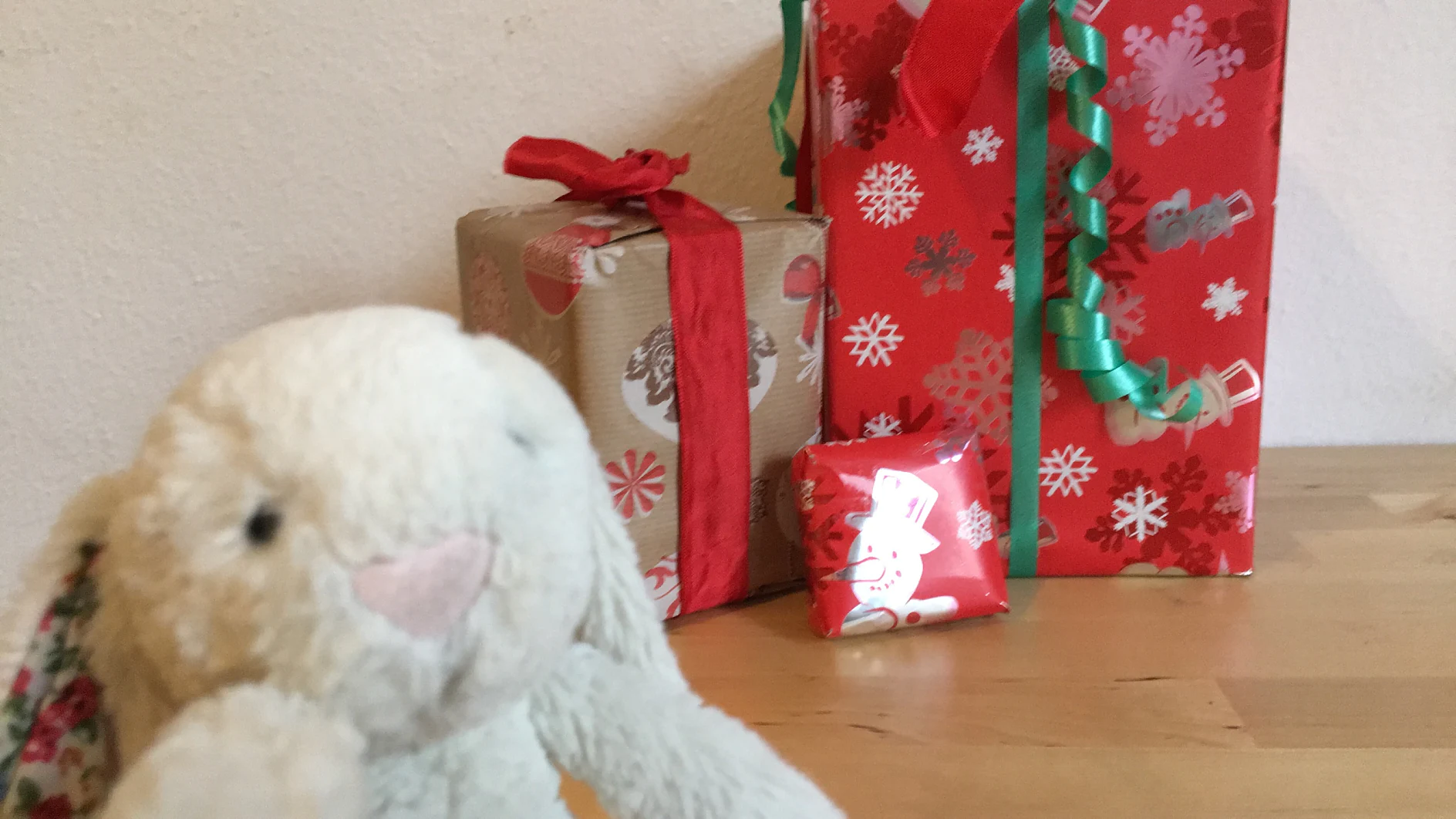 Im Vordergrund steht ein Plüschhase. Im Hintergrund sind drei Geschenke verpackt. Zwei sind in rotem und eines in braunem Papier, alle mit Tesa-Klebeband befestigt und mit roten oder grünen Bändern mit Schneeflockenmuster geschmückt. Sie liegen auf einer Holzfläche. (This text has been generated by AI)