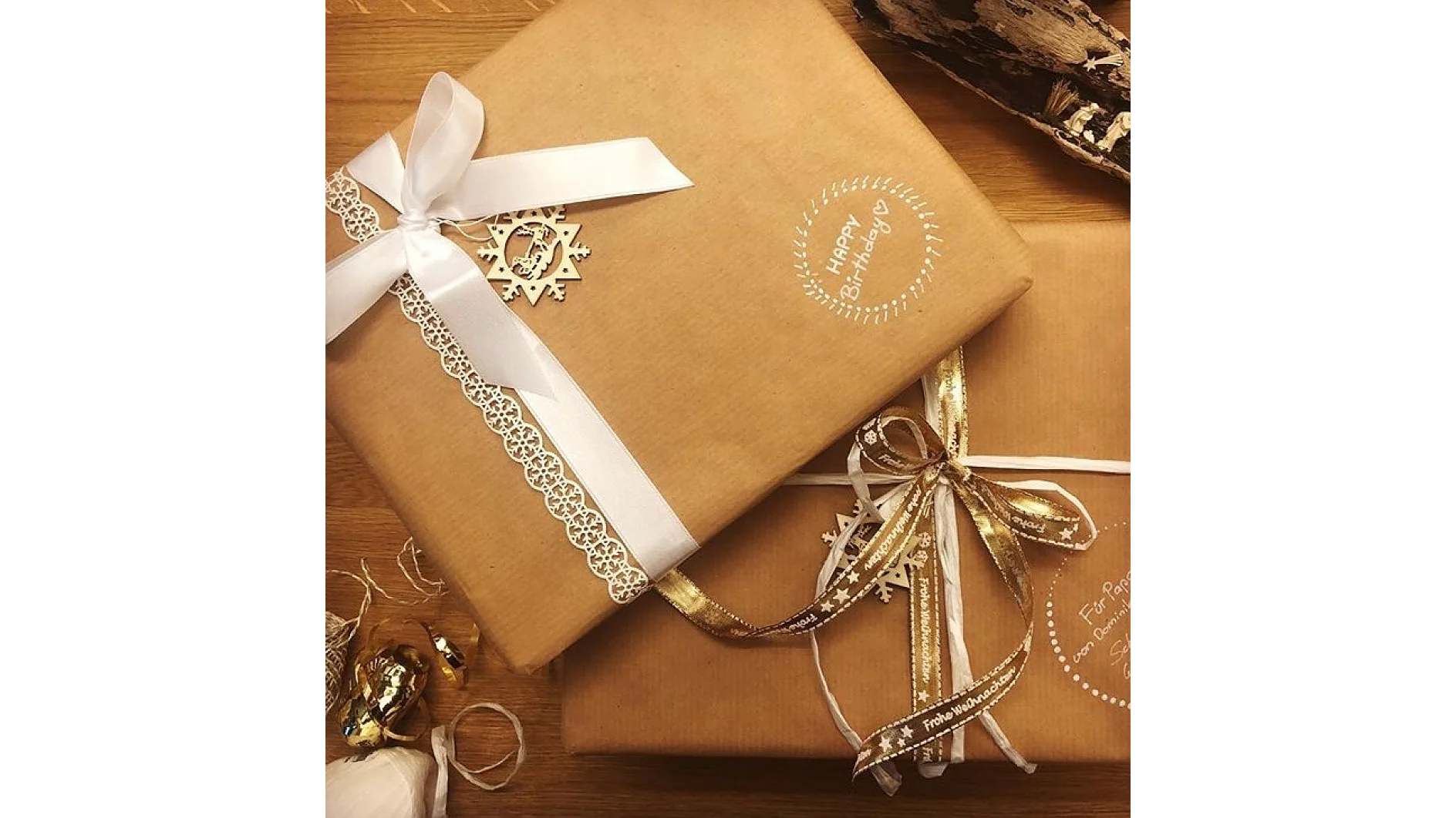 Zwei in braunes Papier eingewickelte Geschenke liegen auf einer Holzoberfläche. Eines ist mit einer weißen Schleife und Spitze geschmückt und trägt einen Schneeflockenanhänger und den Text „Happy Birthday“. Das andere hat eine goldene Schleife und ein Schild in deutscher Sprache. Daneben liegen weiße Schleife, Papier und Tesa-Klebeband. (This text has been generated by AI)