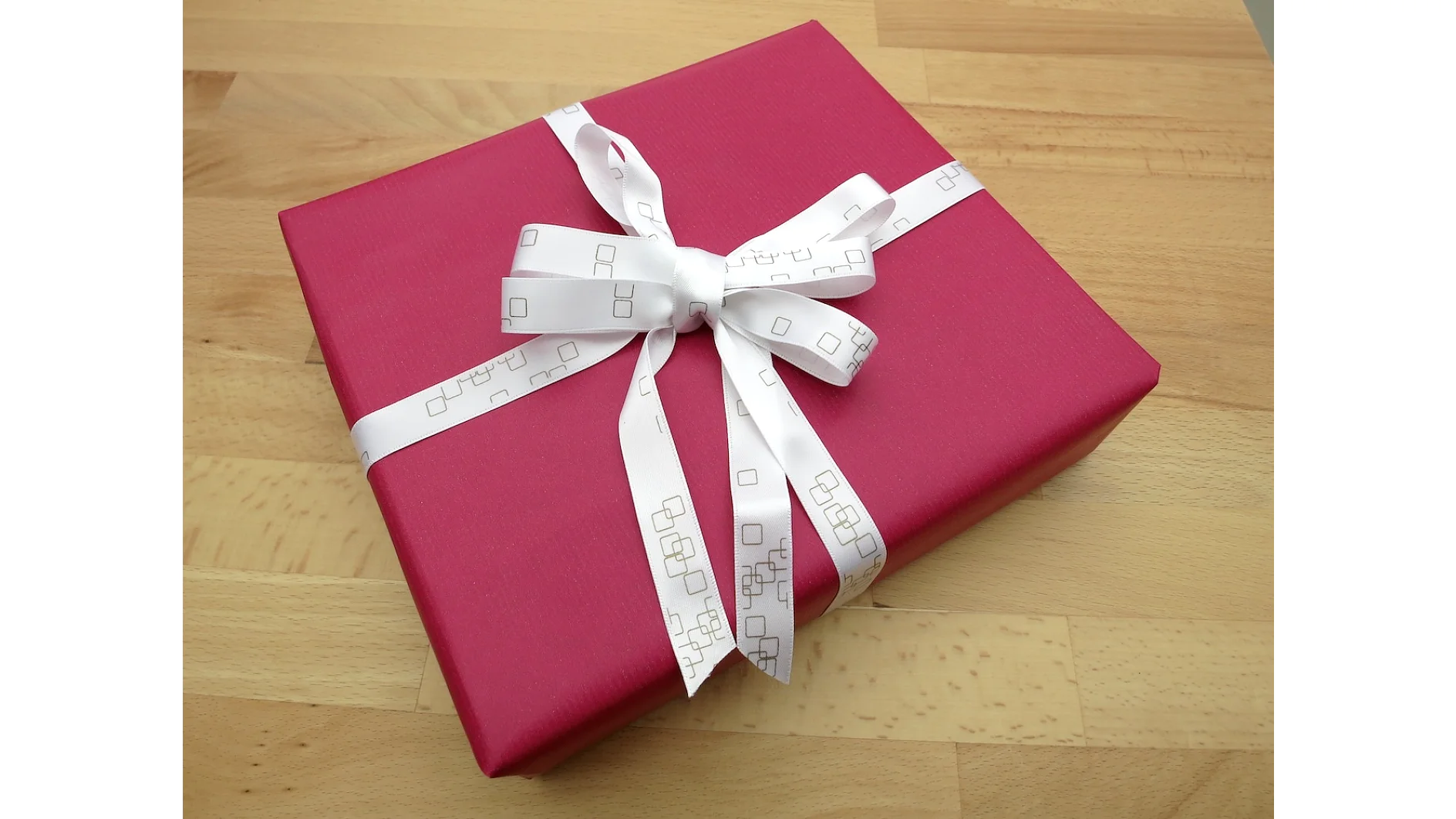 Auf einer Holzoberfläche liegt eine hübsch verpackte Geschenkschachtel mit dunkelrosa Geschenkpapier. Sie ist mit einer weißen Schleife mit einem Muster aus kleinen Quadraten verschnürt und mit Tesa-Klebeband perfekt befestigt. (This text has been generated by AI)