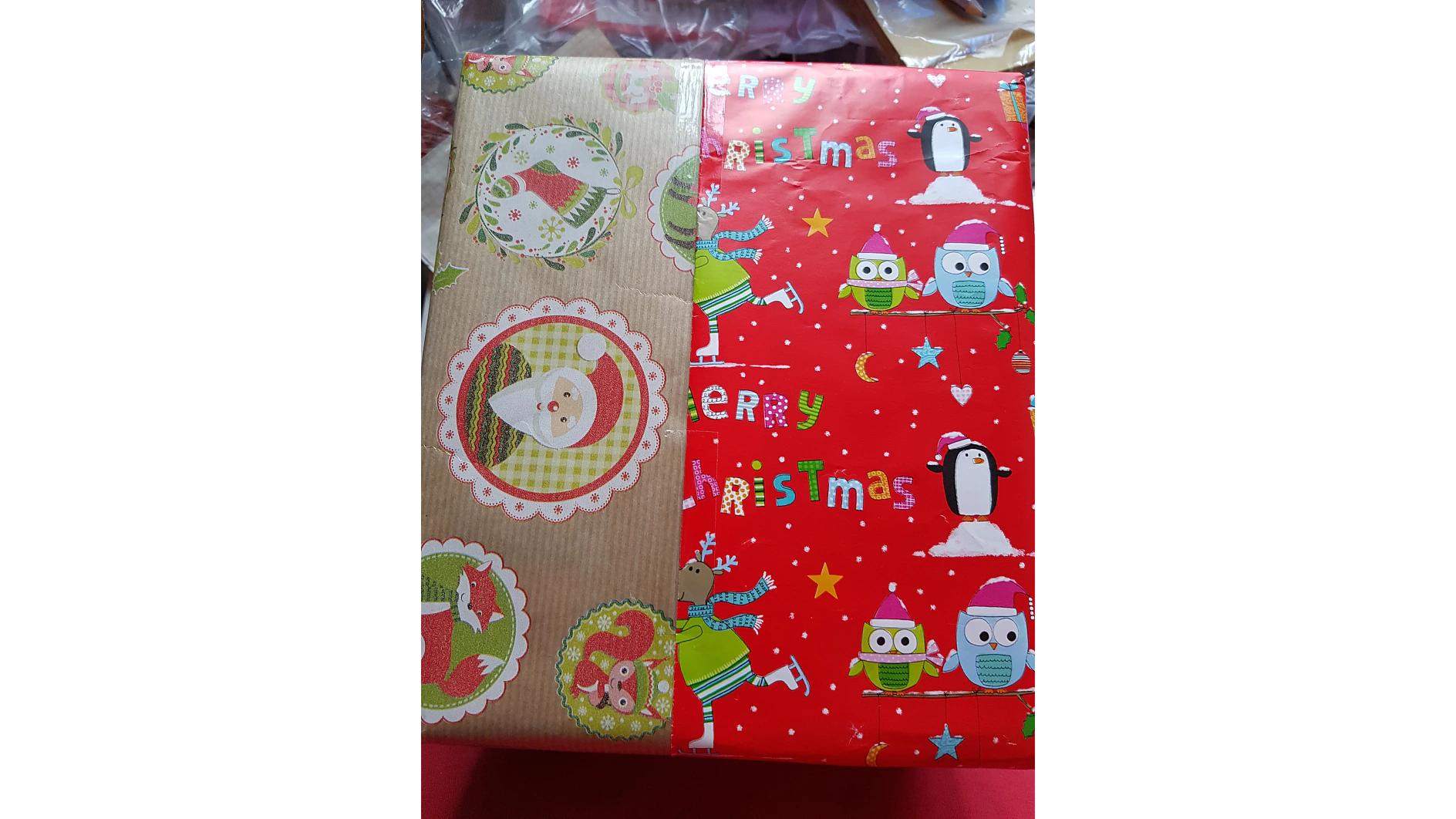 Ein rechteckiges Geschenk, das mit zwei Arten weihnachtlichen Papiers verpackt ist. Eine Seite hat ein braunes Design mit dem Weihnachtsmann und Tieren. Die andere Seite ist rot mit „Frohe Weihnachten“, Pinguinen und Eulen mit festlichen Hüten, alles sorgfältig mit Tesa-Klebeband befestigt. (This text has been generated by AI)