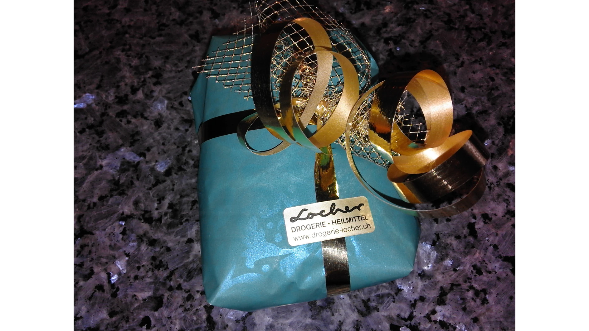 Ein kleines Geschenk, eingewickelt in türkisfarbenes Papier mit einer goldenen Schleife und einer Maschenschleife obendrauf, platziert auf einer gesprenkelten Granitoberfläche. Die Verpackung ist mit Tesaband befestigt. Ein Etikett zeigt den Text „Drogerie – Heilmittel www.drogerie-locher.ch“. (This text has been generated by AI)