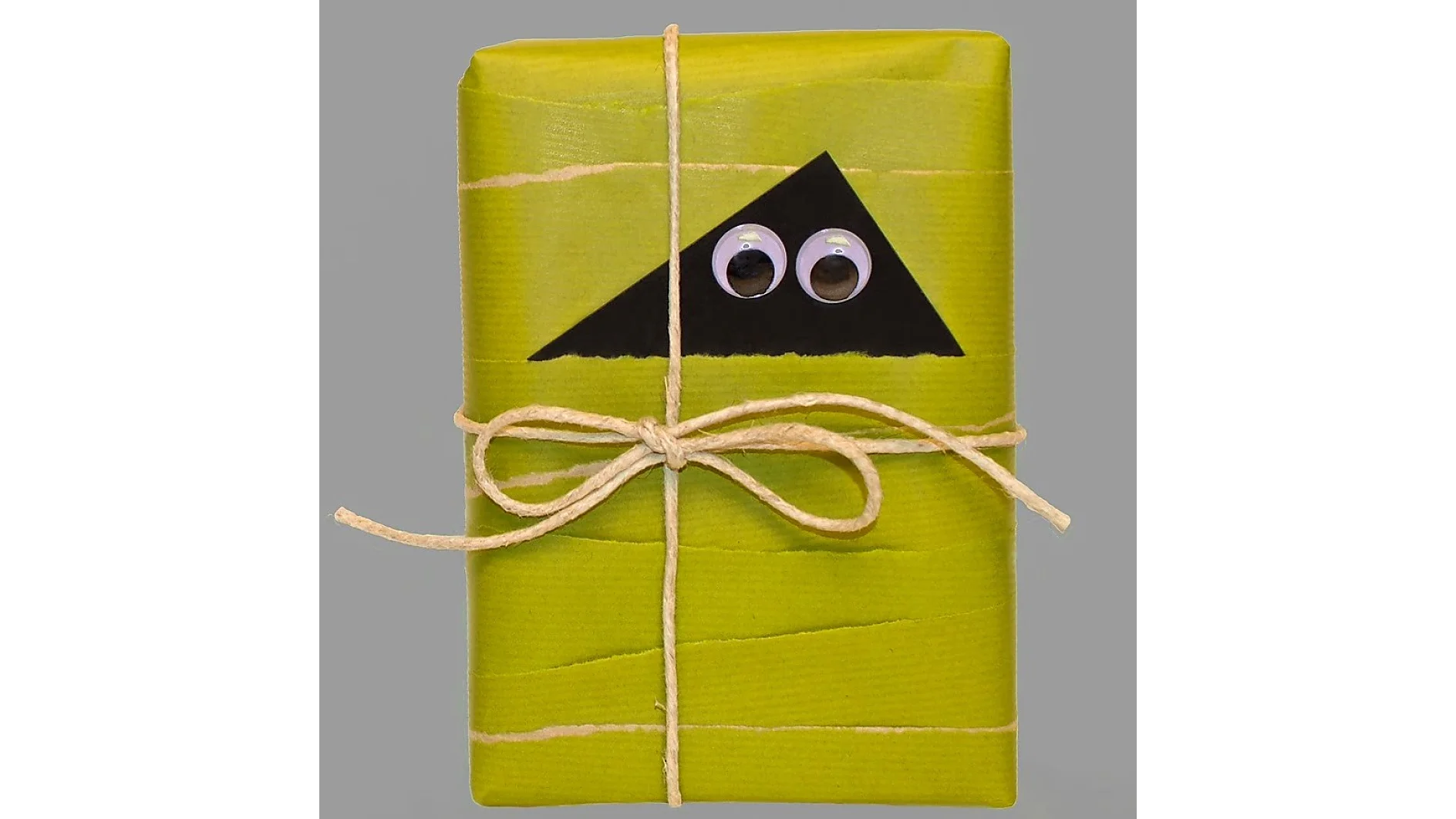 Ein Geschenk, eingewickelt in grünes Papier mit rauer Struktur, mit schwarzem Dreieck und zwei Wackelaugen. Es ist mit einer einfachen Schleife aus Juteschnur zusammengebunden und mit Tesaband zusammengehalten. (This text has been generated by AI)