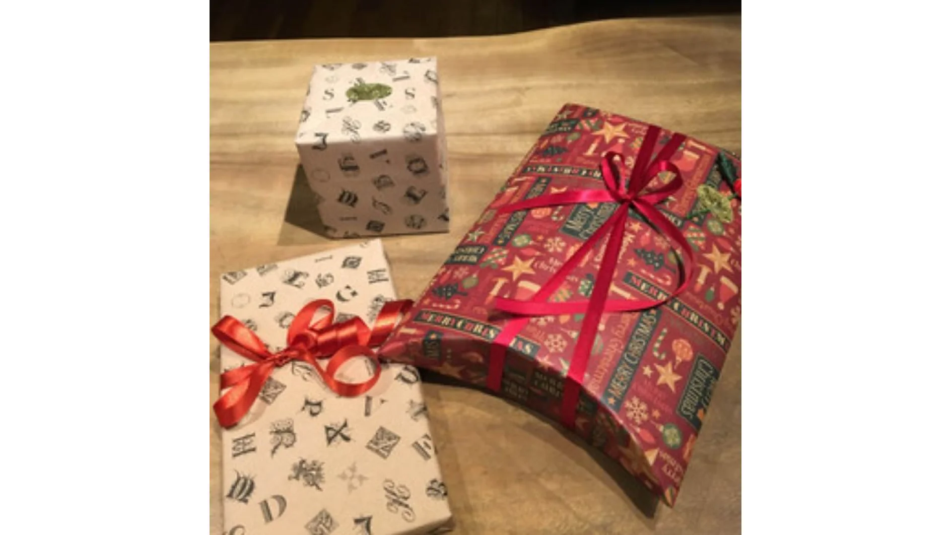 Drei verpackte Geschenke liegen auf einer Holzoberfläche. Das linke Geschenk ist rechteckig und besteht aus cremefarbenem Papier und einer roten Schleife, die mit Tesaband befestigt ist. Das hintere Geschenk ist aus weißem Papier gewürfelt, mit Tesaband zusammengehalten und mit einer grünen Schleife geschmückt. Das rechte Geschenk ist rechteckig und besteht aus rotem gemustertem Papier, das hübsch mit Tesaband befestigt und mit einer roten Schleife zusammengebunden ist. (This text has been generated by AI)