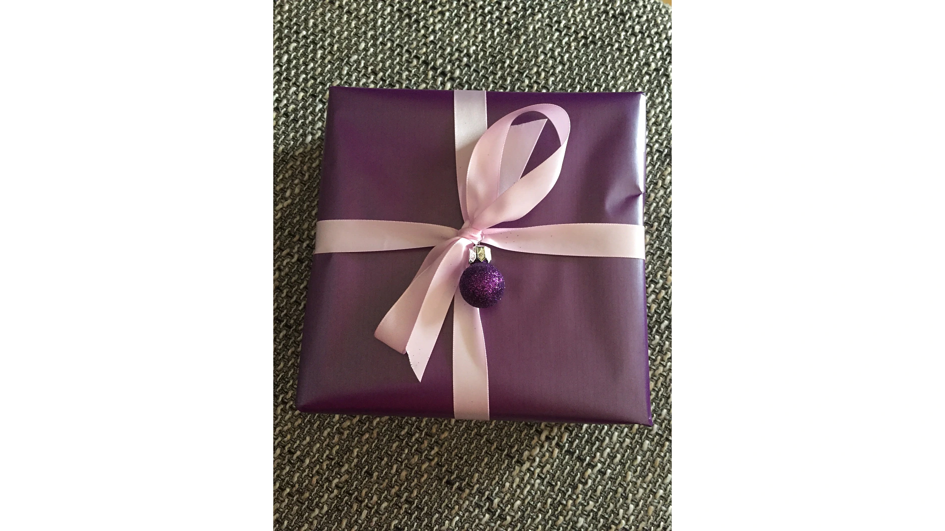 Ein quadratisches Geschenk, eingewickelt in lila Papier mit einer hellrosa Schleife. An der Schleife hängt ein kleines lila Glitzerornament. Das Paket ist mit Tesa-Klebeband befestigt und liegt auf einer strukturierten grauen Oberfläche. (This text has been generated by AI)