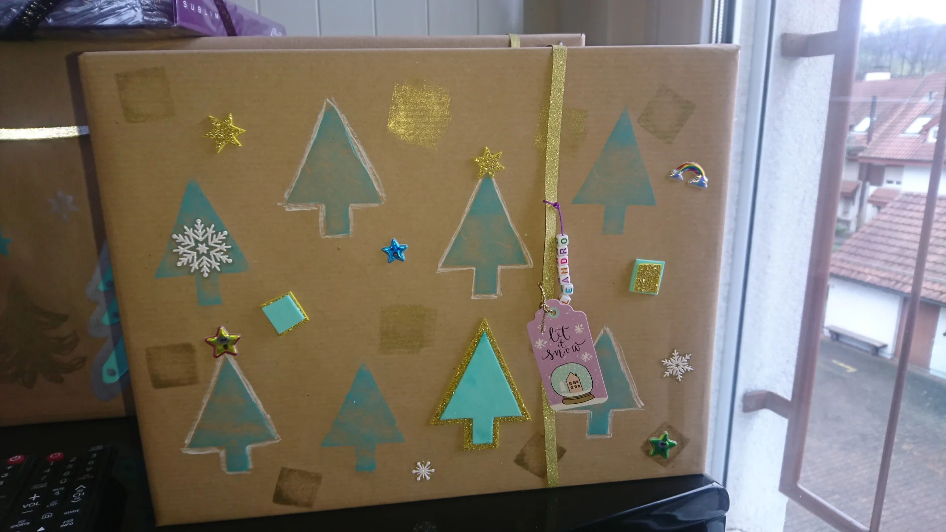 Eine große braune Geschenkbox, verziert mit grünen Baumformen, goldenen Quadraten und Sternaufklebern. Ein goldenes Band verläuft vertikal mit einem Schild, auf dem „Überraschung!“ steht. Die Box steht neben einem Fenster mit Blick auf die Dächer draußen, alles wird sicher mit Tesa-Klebeband zusammengehalten. (This text has been generated by AI)