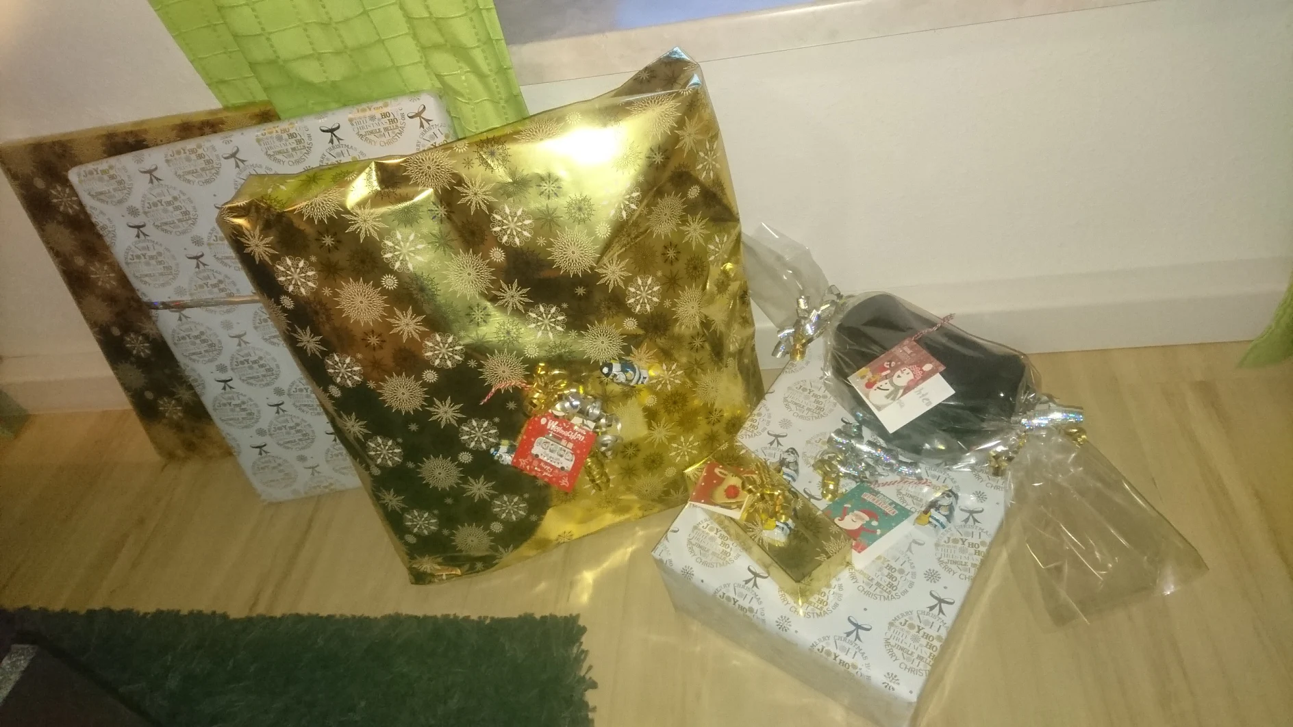 Mehrere verpackte Geschenke liegen auf einem Holzboden neben einem grünen Teppich und einem Vorhang. Die Geschenke sind festlich verpackt, unter anderem in goldenem und weißem Papier mit weihnachtlichen Motiven. An einigen Geschenken sind glänzende Bänder und Anhänger befestigt, alles ordentlich mit Tesa-Klebeband befestigt. (This text has been generated by AI)