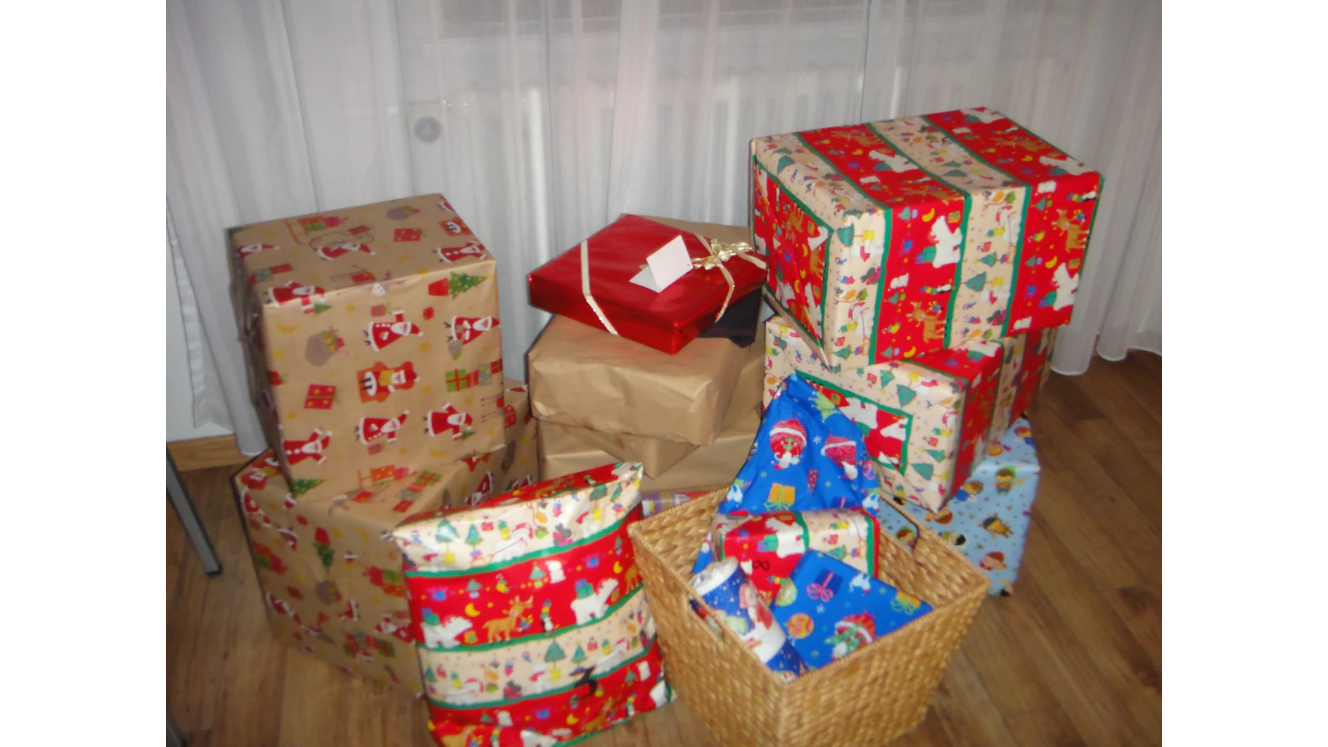 Eine Sammlung in buntes Papier verpackter Geschenke, darunter rote, blaue und mehrfarbige Muster mit festlichen Motiven, auf einem Holzboden platziert. Einige Geschenke sind gestapelt und in einem Korb liegen mehrere verpackte Pakete. Die Verpackung aller Geschenke wurde mit Tesa-Klebeband befestigt. Im Hintergrund ist ein Vorhang zu sehen. (This text has been generated by AI)