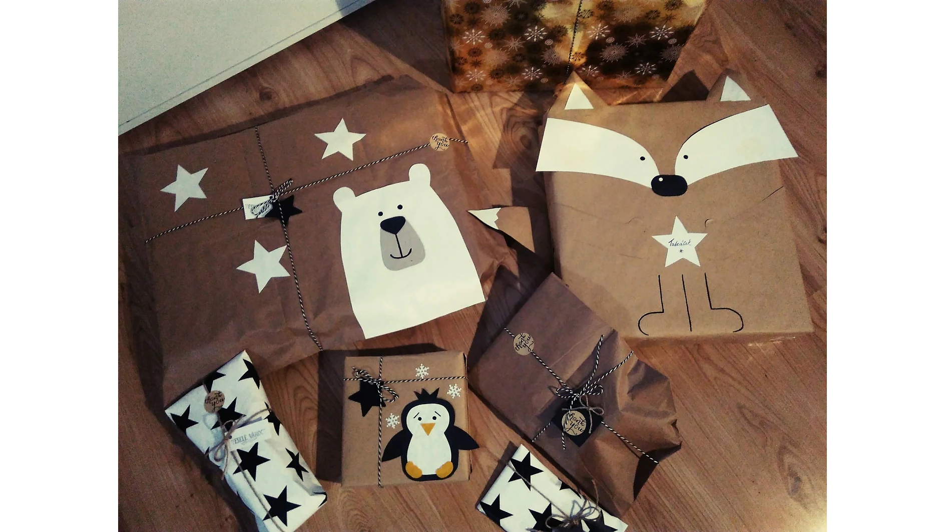 Eine Sammlung verpackter Geschenke auf einem Holzboden. Die Geschenke sind in braunes und weißes Papier eingewickelt und zeigen Motive von Eisbären, Pinguinen und Füchsen. Zur Verzierung werden sternförmige Dekorationen und Schnüre verwendet, alles sorgfältig mit Tesa-Klebeband befestigt. (This text has been generated by AI)
