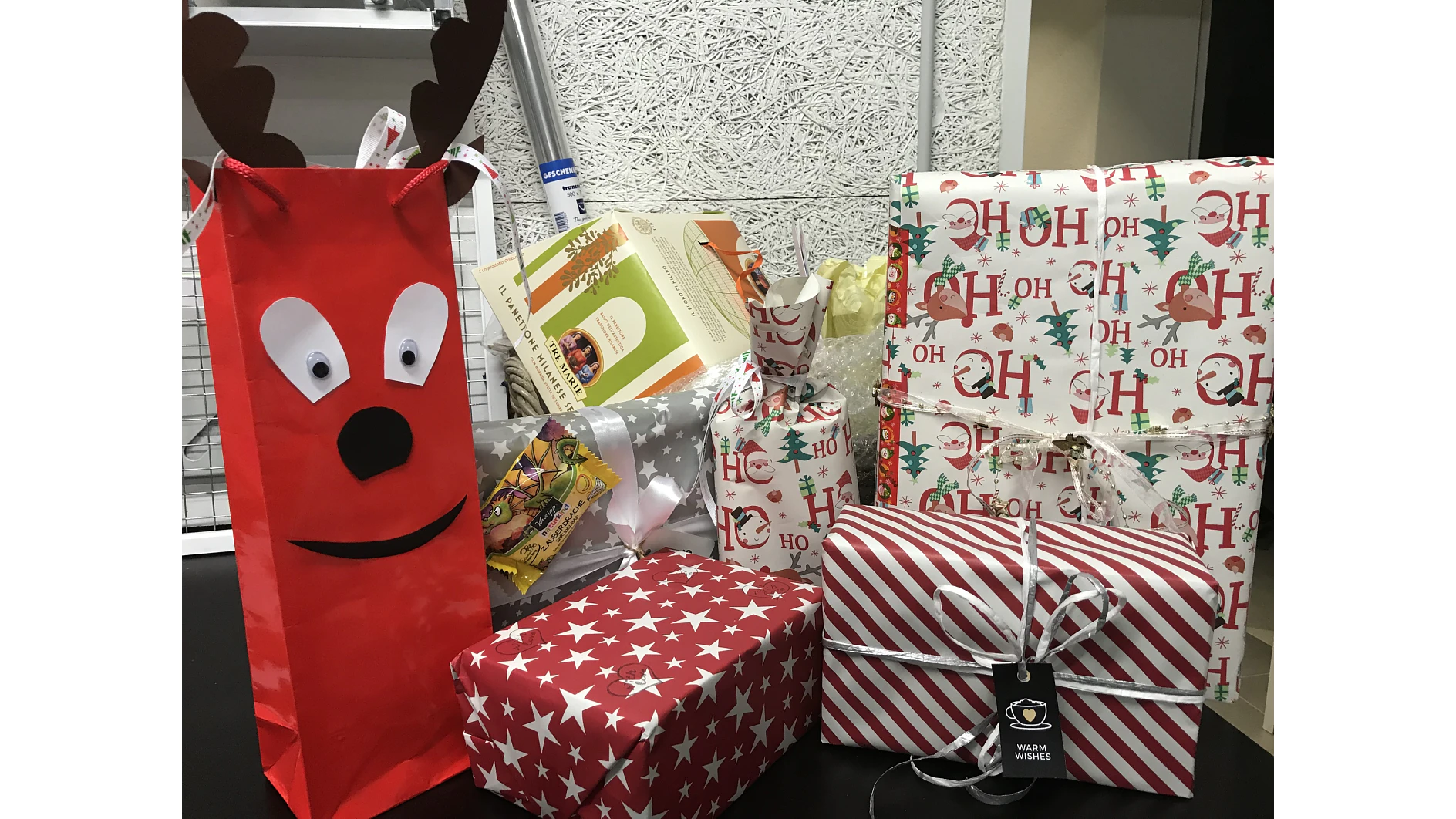 Eine Sammlung von Geschenken mit Feiertagsmotiven wird ausgestellt. Es gibt eine rote Geschenktüte mit Rentiermotiv, mehrere in festliches Papier eingewickelte Geschenke und eine Schleife. Ein Geschenk ist in rotes Sternenpapier eingewickelt, ein anderes ist gestreift. Ein Etikett mit der Aufschrift „Herzliche Grüße“ ist sichtbar. Alle Geschenke sind für ein elegantes Finish sorgfältig mit Tesa-Klebeband befestigt. (This text has been generated by AI)