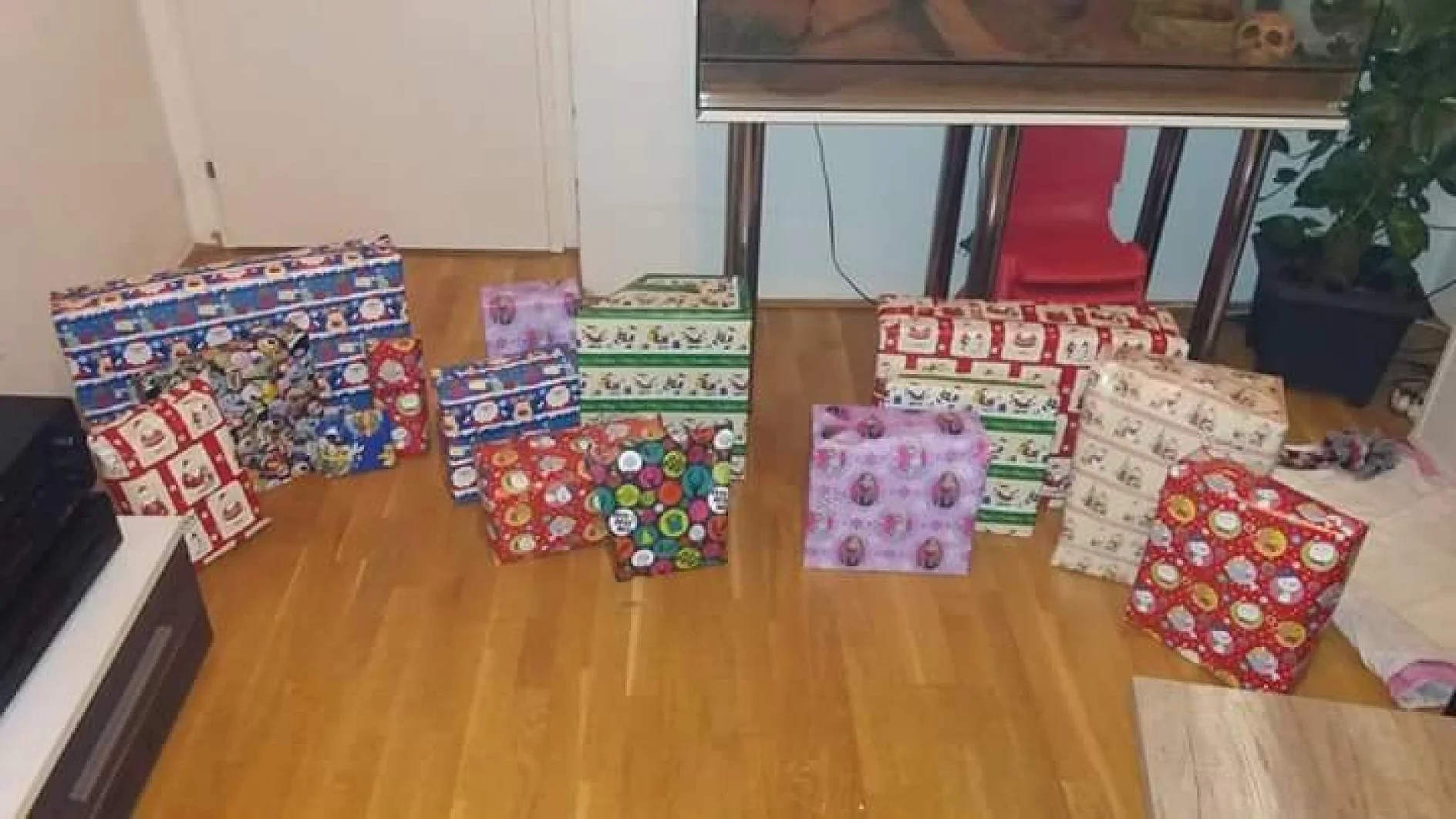 Eine Sammlung unterschiedlich großer und bunter Geschenke, die mit Tesa-Klebeband verpackt sind, liegt auf einem Holzboden. Das Geschenkpapier weist verschiedene festliche Muster auf. Im Hintergrund sind ein Tisch, eine Topfpflanze und ein Teil eines Aquariums zu sehen. (This text has been generated by AI)