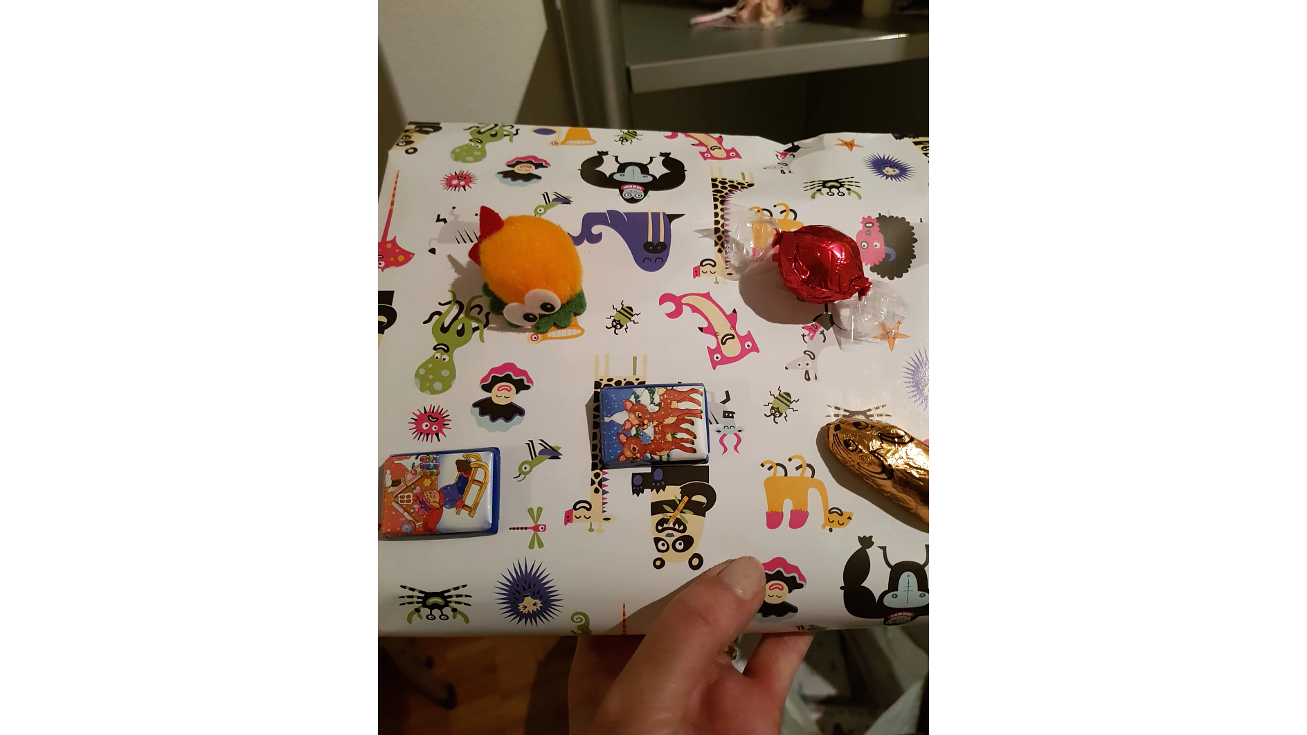 Eine Hand hält ein verpacktes Geschenk mit einem bunten, skurrilen Muster aus abstrakten Tieren. Die Verpackung ist mit Tesa-Klebeband befestigt. Oben liegen ein kleines Plüschtier, zwei eingewickelte Schokoladenstücke und ein Rechteck mit einem Cartoon-Bild. Im Hintergrund ist ein Teil eines Zimmers zu sehen. (This text has been generated by AI)