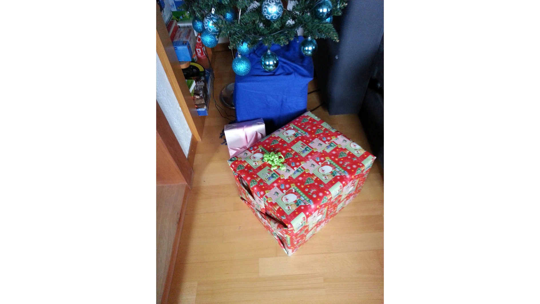 Ein mit blauen Ornamenten geschmückter Weihnachtsbaum ist teilweise sichtbar. Auf dem Boden liegen zwei verpackte Geschenke; eines ist rot mit Weihnachtsmann-Papier und einer grünen Schleife, das andere ist rosa mit einer dunklen Schleife. Der Innenbereich ist mit Holzboden ausgelegt. (This text has been generated by AI)
