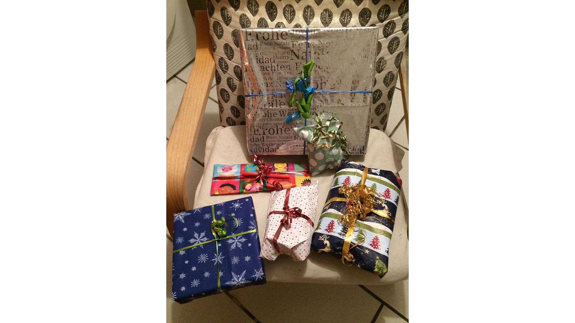 Auf einem gepolsterten Stuhl liegen sechs verpackte Geschenke in unterschiedlichen Größen. Das Geschenkpapier ist mit Tesa-Klebeband befestigt und weist unterschiedliche Motive auf, darunter Schneeflocken, Weihnachtsbäume und festliche Worte. Bänder und Schleifen in verschiedenen Farben zieren die Geschenke. (This text has been generated by AI)