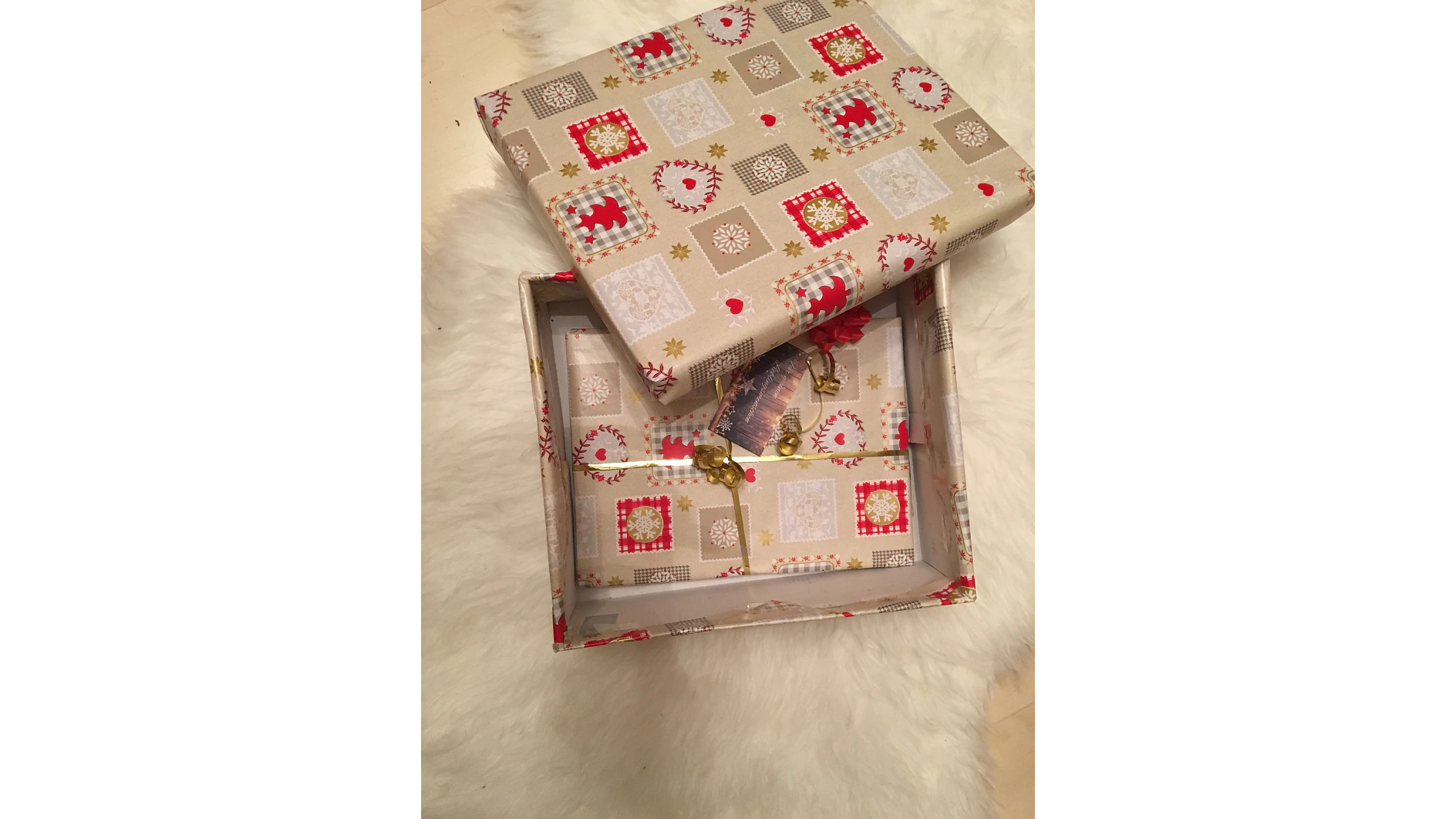 Eine offene Geschenkbox mit einem dekorativen Weihnachtsmuster aus roten und goldenen Symbolen. Im Inneren befinden sich eine kleine Karte und eine goldene Schere, die mit Tesa-Klebeband zusammengehalten werden. Die Box liegt auf einer weißen, flauschigen Oberfläche. (This text has been generated by AI)