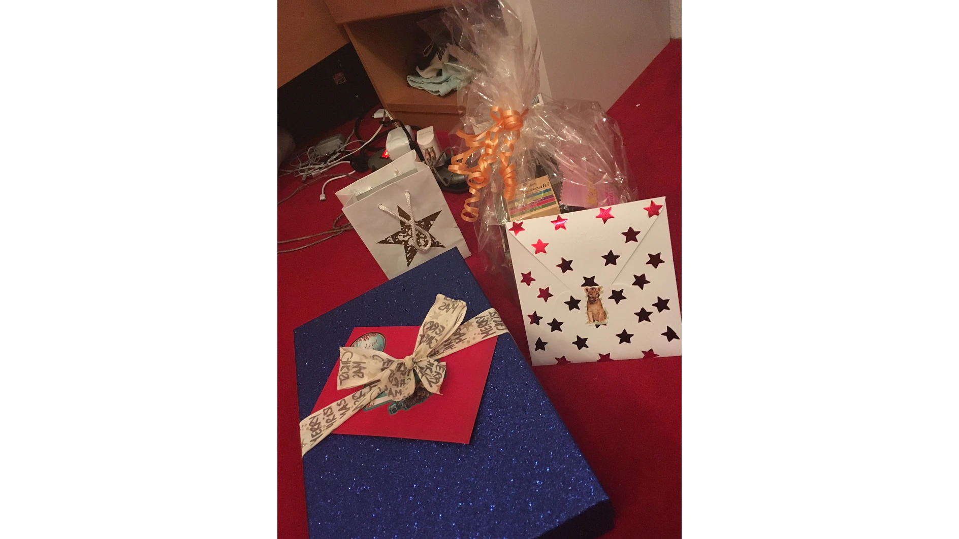 Ein Foto zeigt eine Sammlung von Geschenkpaketen auf einem roten Untergrund. Eines davon ist eine blaue Glitzerschachtel, ein anderes ein weißer Umschlag mit Sternenmuster und einer Hundeillustration, ein kleiner verzierter weißer Beutel und ein in Zellophan verpackter Artikel mit einer orangefarbenen Schleife, die mit Tesaband befestigt ist. (This text has been generated by AI)