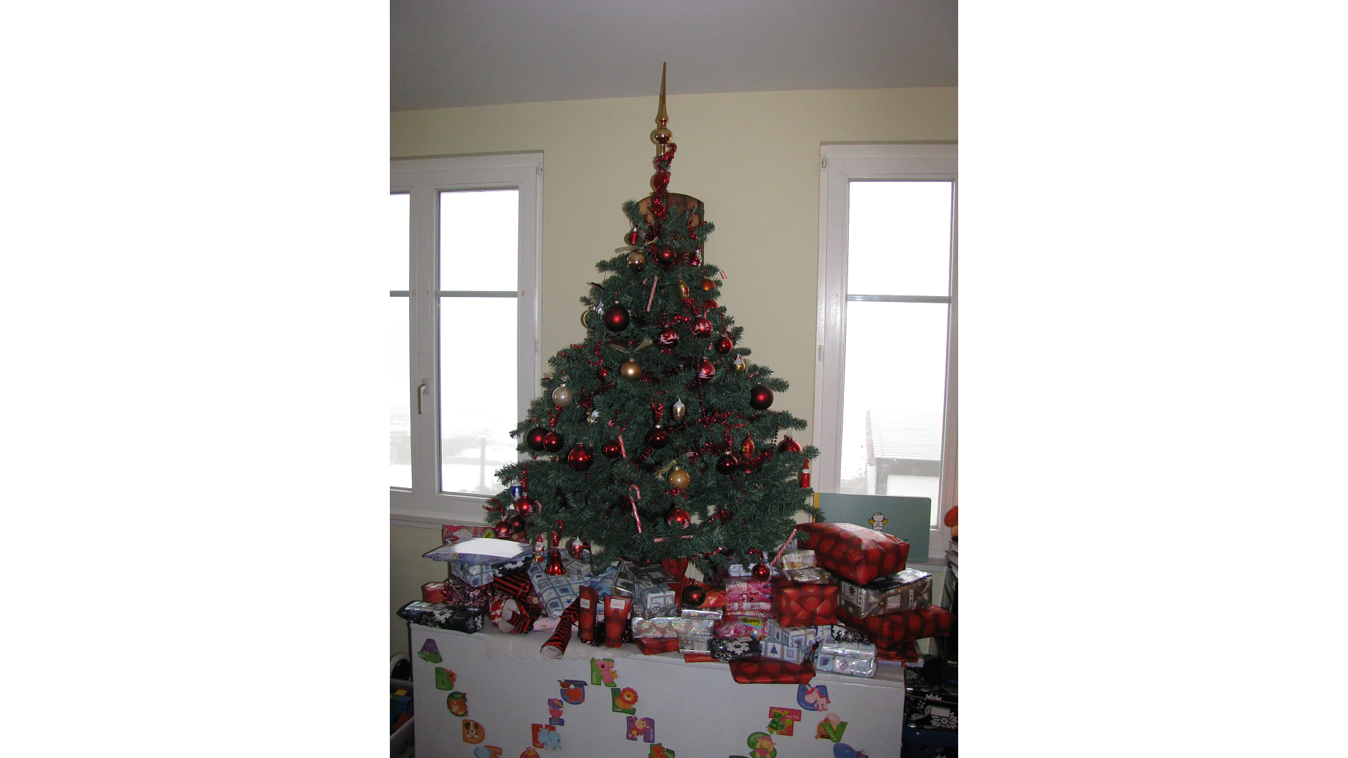 Drinnen neben zwei Fenstern steht ein geschmückter Weihnachtsbaum, geschmückt mit roten und goldenen Ornamenten und einer goldenen Spitze. Unter und um den Baum herum sind zahlreiche in rotem, silbernem und gemustertem Papier verpackte Geschenke platziert. (This text has been generated by AI)