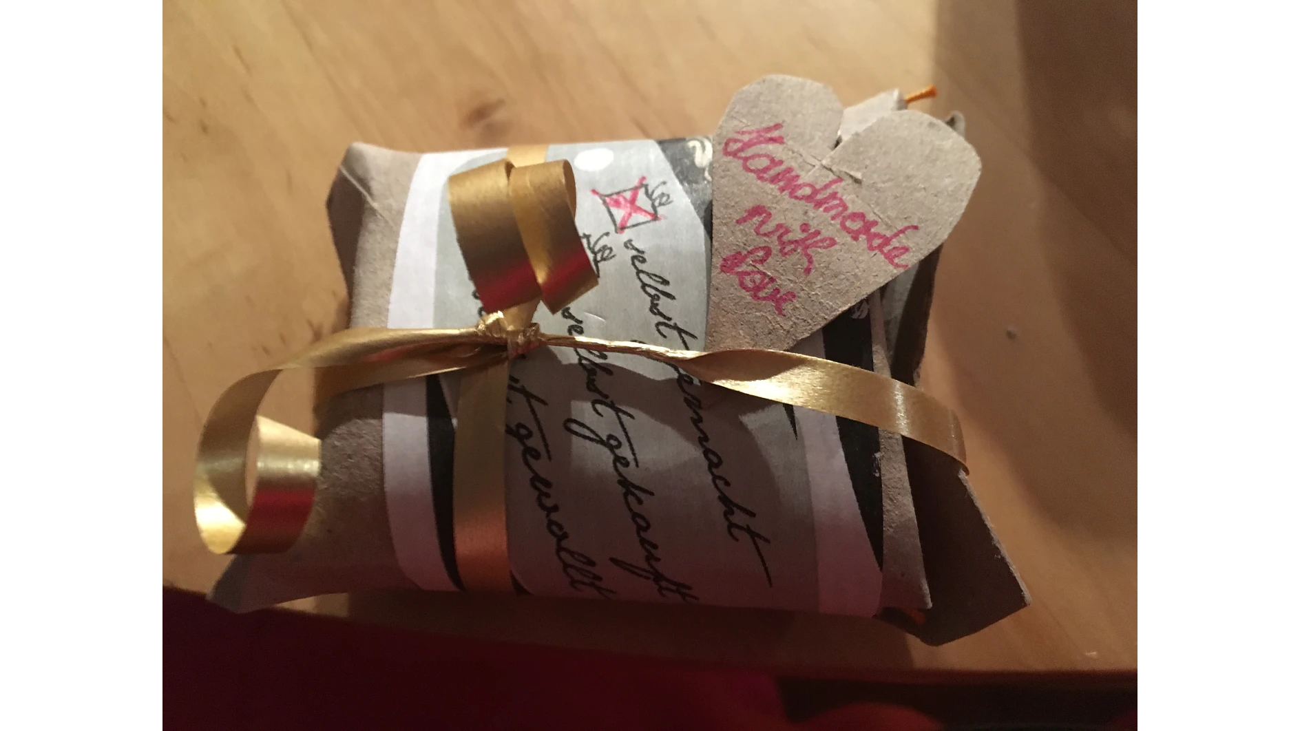 Abgebildet ist ein kleines Geschenk, das in braunes Papier eingewickelt und mit einer goldenen Schleife befestigt ist. Auf einem herzförmigen Etikett am Geschenk steht in rosa und schwarzer Tinte „Handmade mit love“. Der Hintergrund ist eine Holzoberfläche. (This text has been generated by AI)