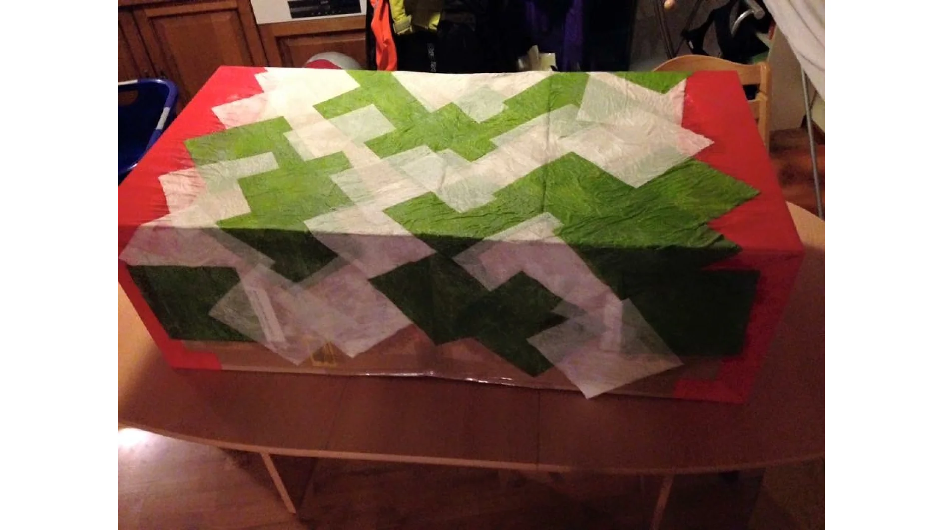 Ein rechteckiges Objekt auf einem Holztisch ist mit rotem, grünem und weißem Strukturpapier bedeckt, das in überlappenden Mustern angeordnet ist. Im Hintergrund ist ein Teil einer Küche mit Schränken zu sehen, und links ist teilweise ein blauer Behälter zu sehen. (This text has been generated by AI)