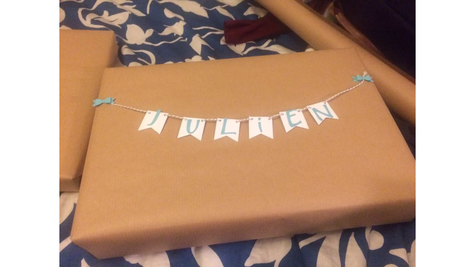 Auf einer Oberfläche liegt ein in braunes Papier eingewickeltes Geschenk, auf dem ein kleines Banner aus Schnur hängt. Auf dem Banner steht in blauen Buchstaben „JULIEN“ auf weißen dreieckigen Fahnen. An jedem Ende des Banners befinden sich blaue Schleifendekorationen, alles sorgfältig mit Tesa-Klebeband befestigt. (This text has been generated by AI)