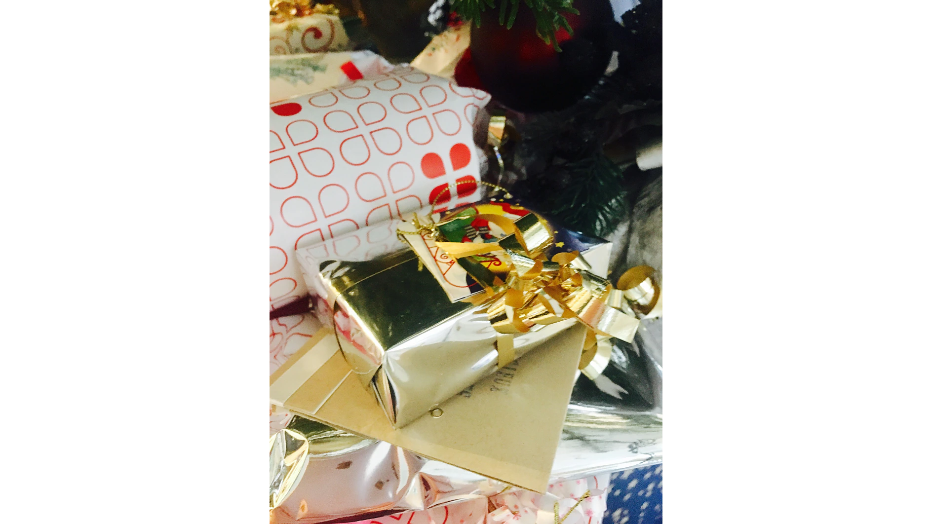 Eine Sammlung verpackter Geschenke ist gestapelt und mit glänzendem goldenen, weißen und gemusterten Geschenkpapier versehen. Einige haben goldene, gekräuselte Bänder. Unter der glänzenden goldenen Schachtel ist ein Umschlag versteckt. In der Ecke ist ein angedeuteter Tannenzweig zu sehen. Die Verpackung ist fachmännisch mit Tesa-Klebeband befestigt, um ein ordentliches und elegantes Aussehen zu erzielen. (This text has been generated by AI)
