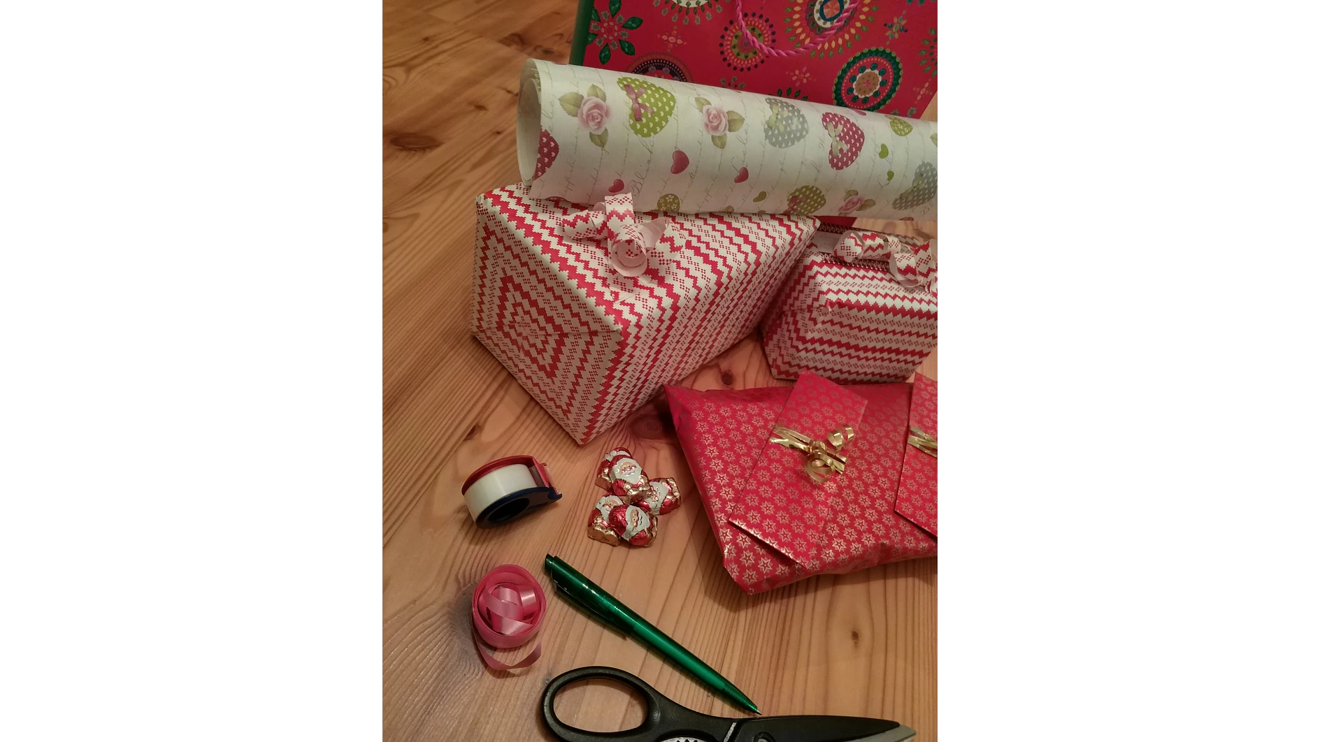 Geschenkverpackungszubehör auf einem Holzboden, darunter drei verpackte Geschenke mit rot-weißem Muster, dekorative Bänder, eine Rolle Geschenkpapier mit Blumenmuster, eine Schere, ein Stift und eine Rolle Tesaband. (This text has been generated by AI)