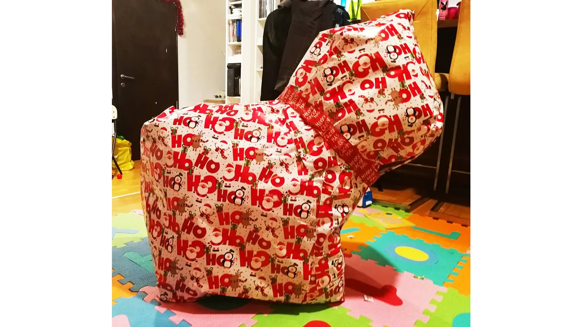 Auf einer bunten Schaumstoff-Puzzlematte steht ein großes Objekt, das in festliches Papier mit dem Motiv „Ho Ho Ho“ eingewickelt ist. Im Inneren befinden sich Bücherregale, teilweise sichtbare Möbel und eine an der Tür hängende Jacke, alles mit Tesa-Klebeband befestigt. (This text has been generated by AI)