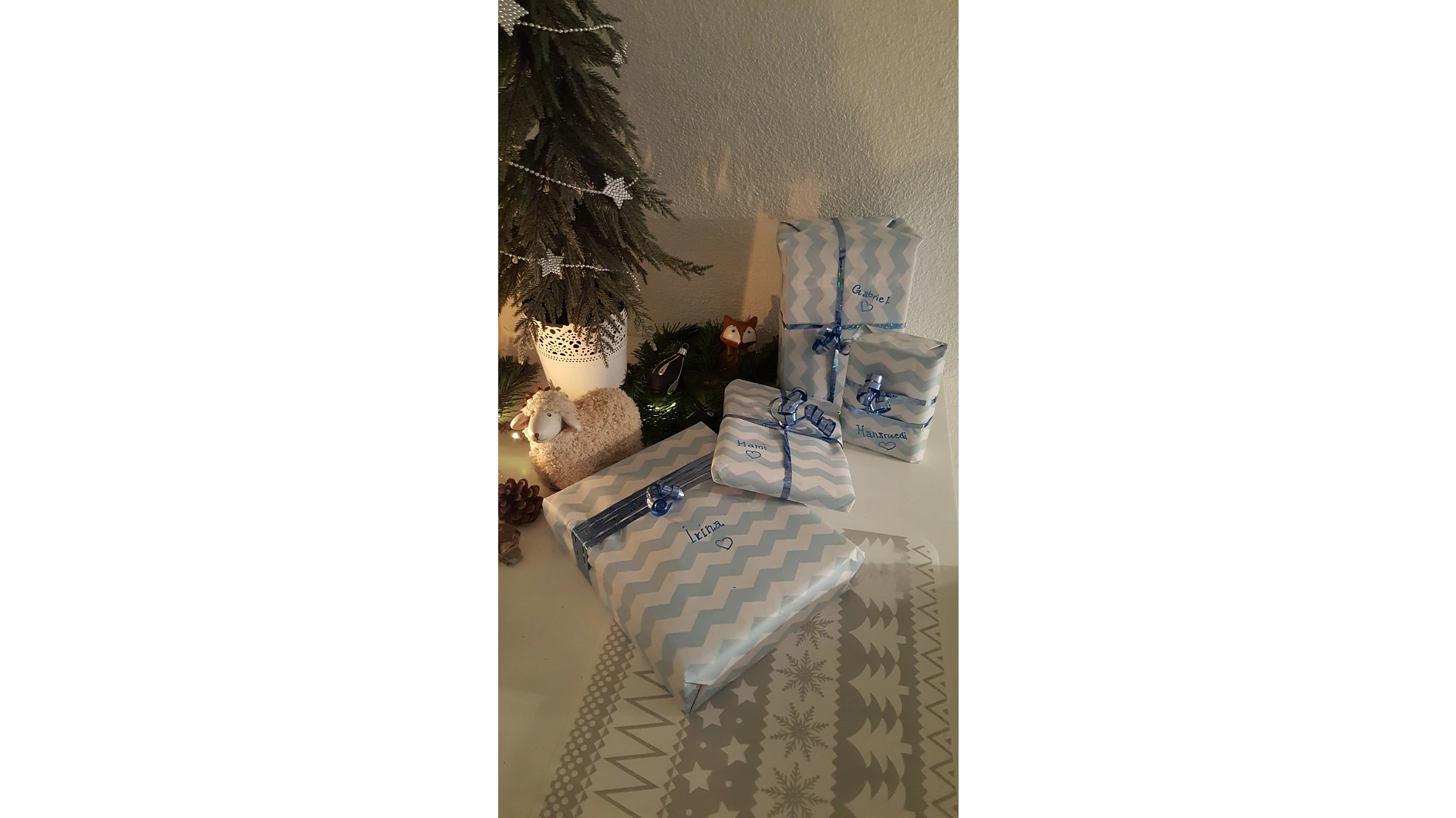 Auf einem Tisch steht ein kleiner geschmückter Baum mit einem festlichen Kranz neben einer Schafsfigur und mit blauem Tesaband verpackten Geschenken. Auf den Geschenken sind handgeschriebene Namen. Neben dem Baum lugt eine kleine Fuchsfigur aus dem Grün hervor. (This text has been generated by AI)